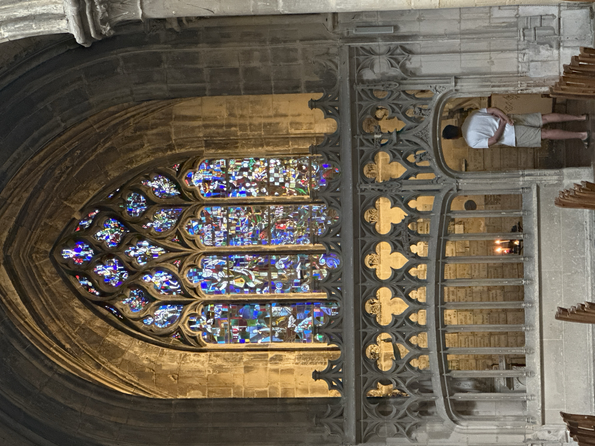 A man stands in a stone archway inside a church, facing away from the camera. He is wearing a white t-shirt and khaki shorts. Behind him, through the archway, is a wall lined with plaques and two lit candles on a ledge. Above the archway is a large stained-glass window with intricate patterns and colorful depictions. The overall environment suggests an old, ornate church interior. The church is the Saint-Jacques church in Dieppe, France.