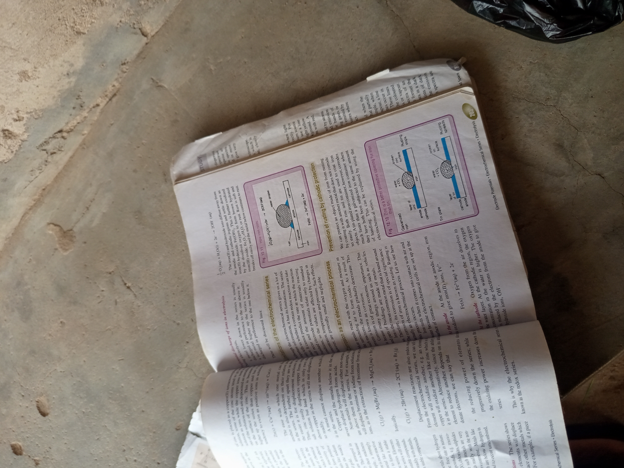 The image shows an open textbook on a textured, concrete-like surface. The textbook pages display text and diagrams related to electrochemistry, including sections on displacement reactions, corrosion, and cathodic protection. One diagram illustrates the process of rusting, with labels for anode, cathode, water droplet, and zinc. Another diagram compares galvanized iron and tin plate in the context of rusting. The text is primarily in English, with some scientific equations and chemical formulas. The overall scene appears to be an indoor setting, possibly a classroom or study area, given the presence of the textbook and the plain background. There are no people or obvious signs of specific location within a city.