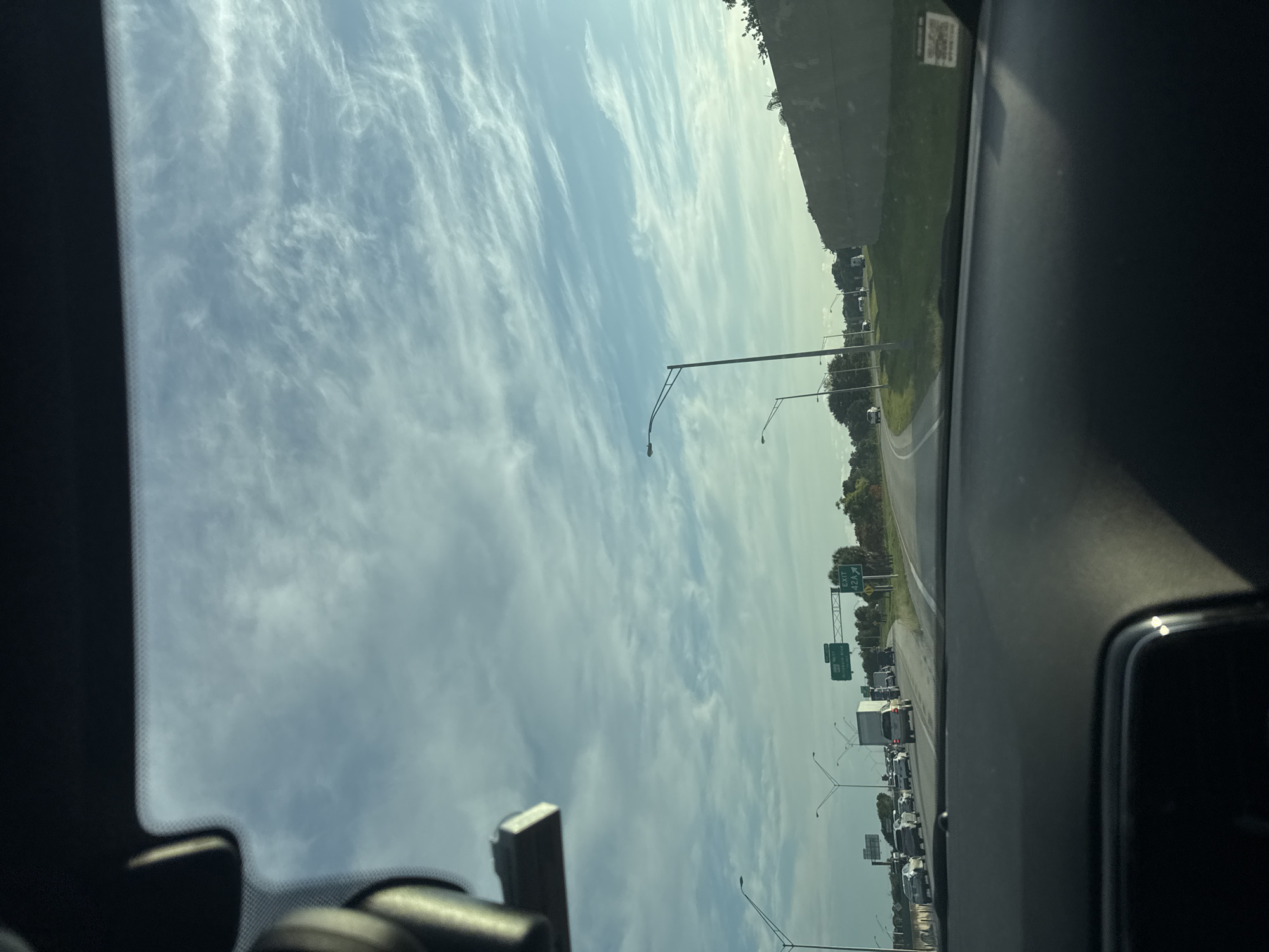 The image shows a view from inside a vehicle, looking out at a highway in Deerfield Beach, Florida, USA. The sky is partly cloudy with wispy cirrus clouds. Traffic is visible on the highway, with several cars and trucks. There are also highway signs indicating an exit for Hillsboro Blvd. The road curves to the right. A concrete barrier is visible on the right side of the highway. The interior of the vehicle, including the dashboard and rearview mirror, is visible in the foreground.