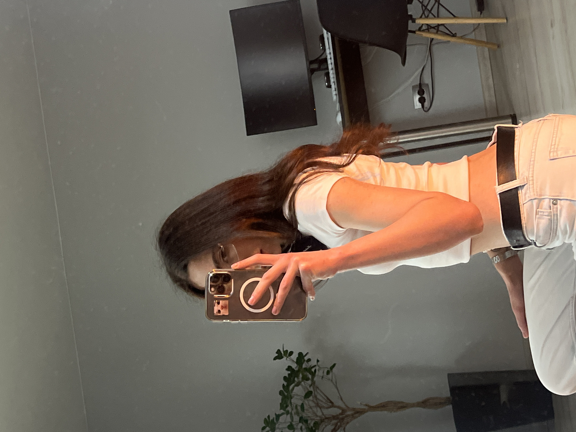 A mirror selfie captures a young woman in an indoor setting in Ternopil, Ukraine. The image is oriented ninety degrees clockwise, showing the woman from the waist up, reflected in a large mirror. She has long, dark brown hair and is wearing a white short-sleeved crop top, light-colored jeans, and a black belt. She holds a smartphone, likely an iPhone with a clear case featuring a white MagSafe ring, to take the picture. Her face is partially obscured by the phone, but one eye and her forehead are visible looking towards the camera. A metallic watch or bracelet is on her left wrist.
The reflected background shows elements of a room, including a dark computer monitor or TV screen, a black office chair with wooden legs, and an electrical outlet with cables plugged into a light-colored wall. A small, green potted plant with branching stems is visible in the lower part of the reflection. The mirror surface displays minor specks, possibly dust. The lighting suggests an indoor environment, but the time of day is not discernible.