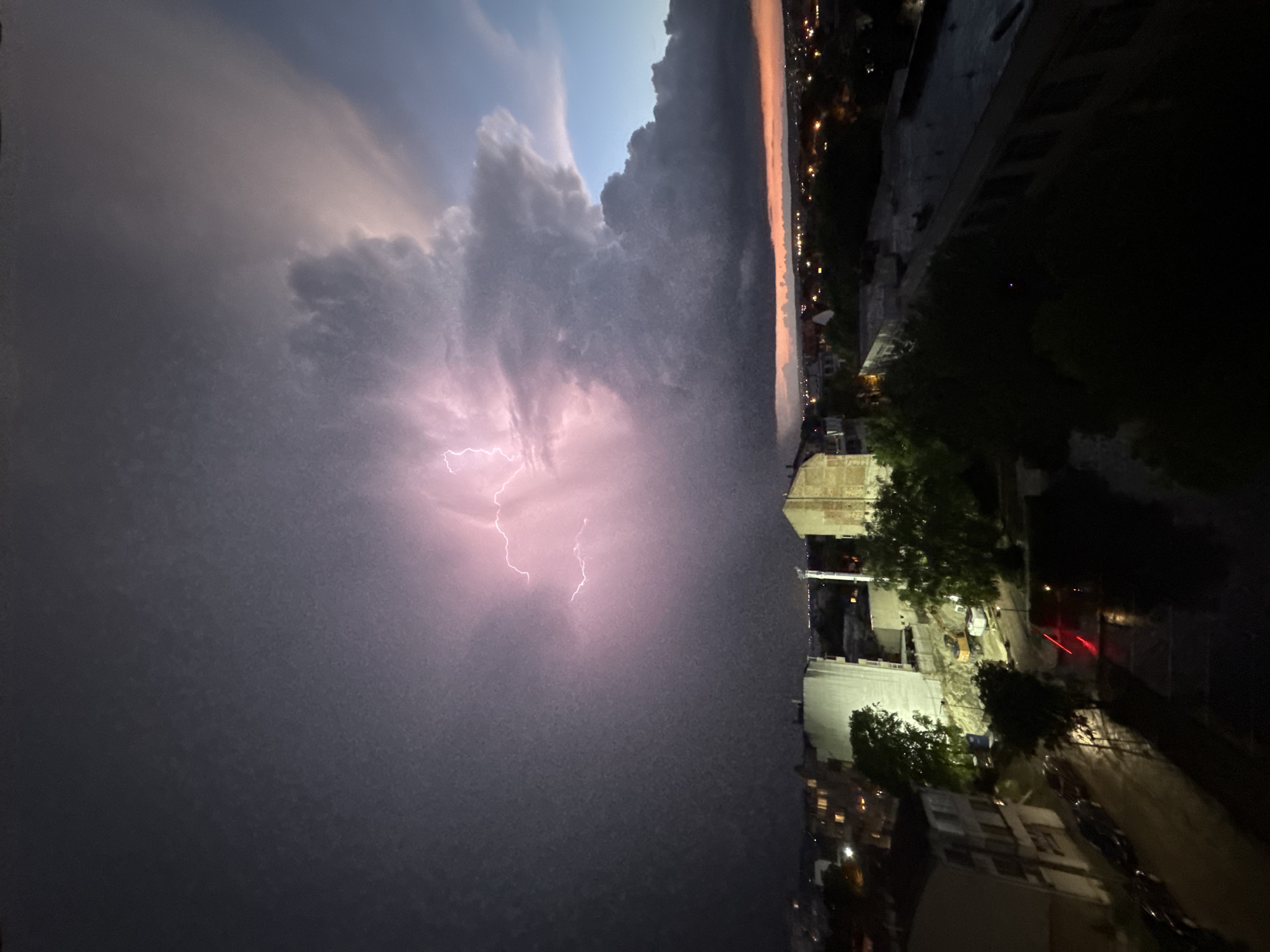 The image shows a thunderstorm over Varna, Bulgaria at night. Lightning is visible within the clouds. The cityscape is partially visible in the foreground. Street lights and car taillights illuminate the dark buildings and roads. The image appears to be taken from a high vantage point, possibly a tall building or hill. The image captures an urban landscape under a stormy sky.