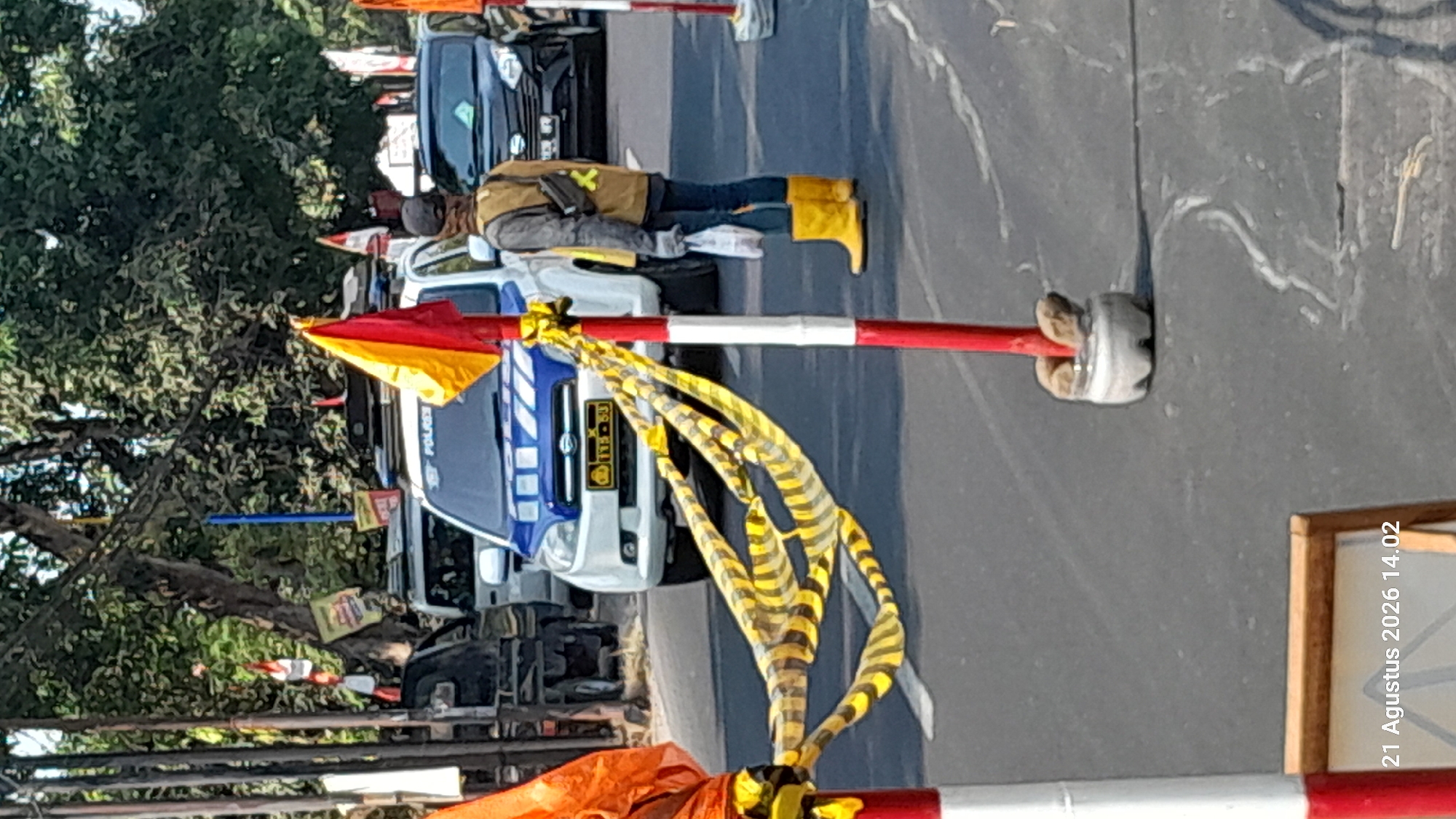 In Takeran, Indonesia, a police vehicle with a blue and white livery is parked on a paved road. A person wearing a brown vest, dark pants, and yellow boots stands to the right of the vehicle, holding a white plastic bag. The vehicle's license plate is visible. Yellow and black caution tape is strung between two red and white striped poles in the foreground, partially obscuring the vehicle and the person. Trees and foliage form the background, with other vehicles also visible on the road. A date and time stamp, "21 Agustus 2026 14.02", is present in the lower left corner of the image.