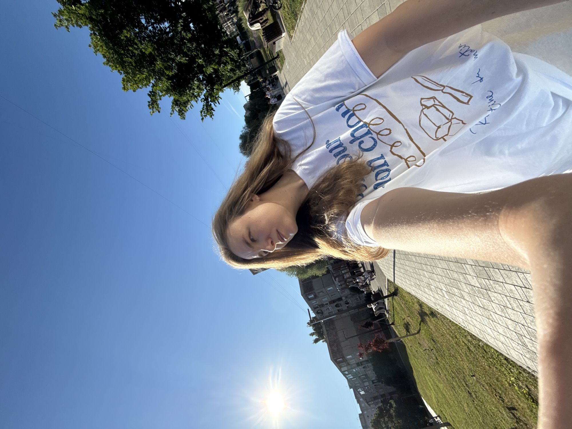 In a wide-angle, low-angle, outdoor selfie, a young woman with long, light brown hair is captured from the chest up, walking along a sun-drenched pedestrian walkway. She is wearing a white t-shirt with blue and brown graphic designs and text that reads "pour mon chou" and "koutine du matin". Her right arm is extended towards the camera.
The background reveals a city scene in Vladimir, Russia, with a clear blue sky, a bright sun beaming from the left, and a line of multi-story apartment buildings. To the right of the walkway, a patch of green grass leads to trees, and further back, other pedestrians are visible, some seated on benches, and a stroller. The walkway is paved with light-colored rectangular tiles arranged in a diamond pattern. The sun casts long shadows across the scene.