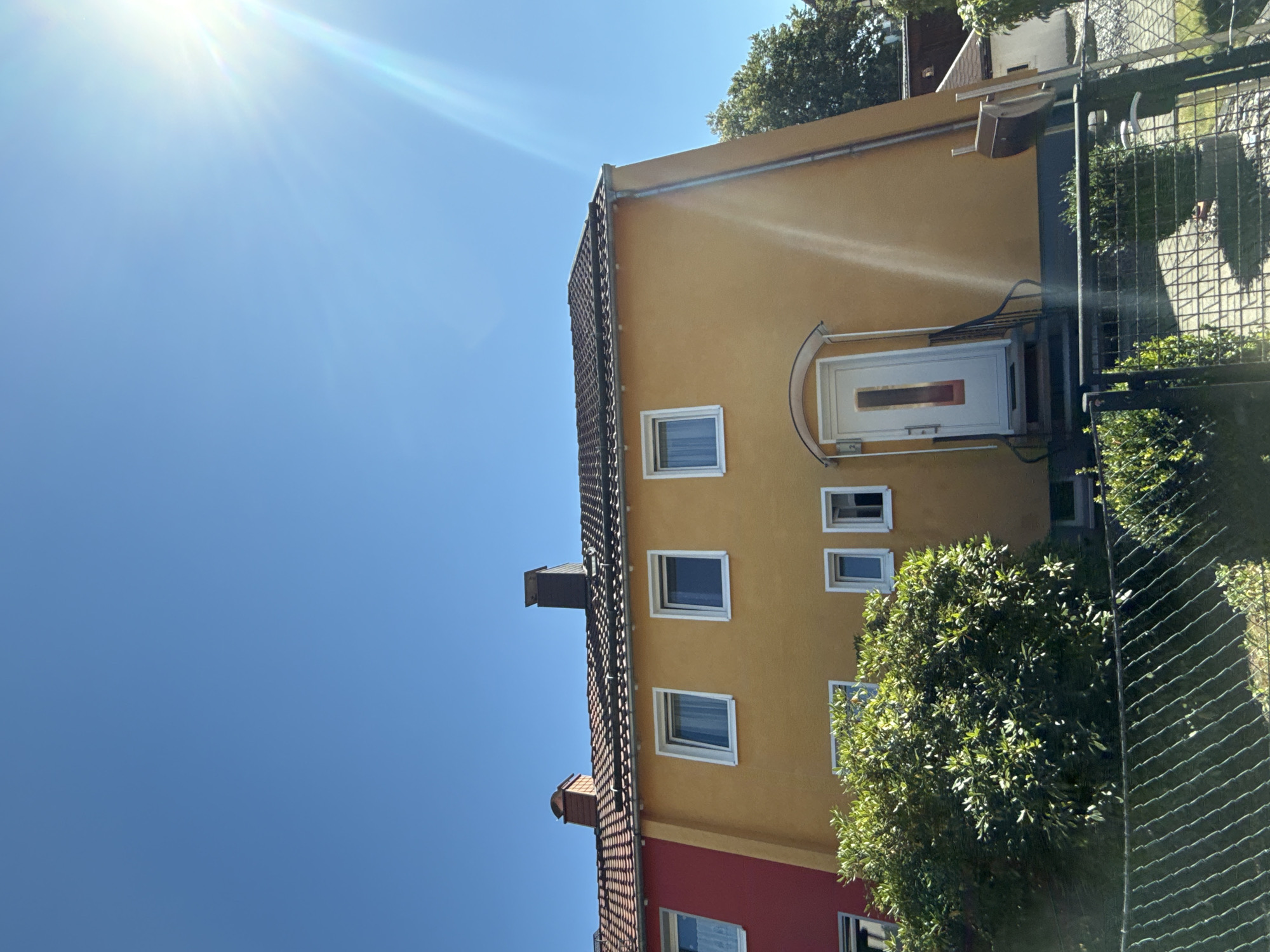 In Bad Sachsa, Germany, on a clear, sunny day, a two-story row house is depicted. The house features a yellow facade, a tiled roof, and several white-framed windows. A red-colored house is partially visible to the left. In front of the yellow house, a white door with a glass pane is set within a small entryway, accessed by a few steps and a metal railing. A lush green bush is situated to the left of the entrance, and a chain-link fence runs across the foreground, partially obscuring the lower part of the scene. The sky is a vibrant blue with the sun prominently visible in the upper right corner, casting lens flares. A chimney with a cap is visible on the roof.