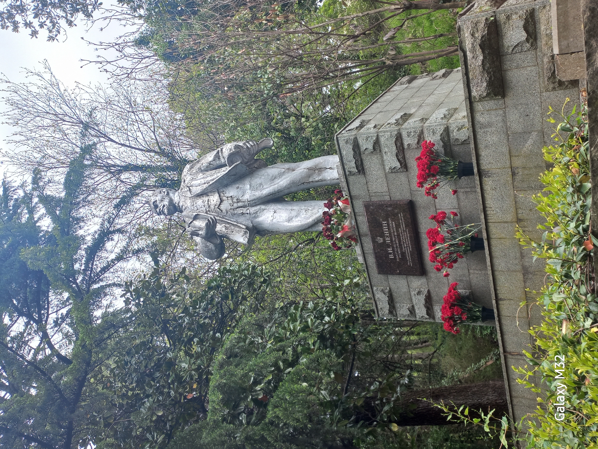 The image shows a silver statue of Vladimir Lenin on a stone pedestal surrounded by trees and foliage. Red carnations are arranged in vases on the pedestal. A plaque bearing an inscription in Russian is mounted on the pedestal. The location is in the city of Sochi, Russia. A watermark "Galaxy M32" is visible.
