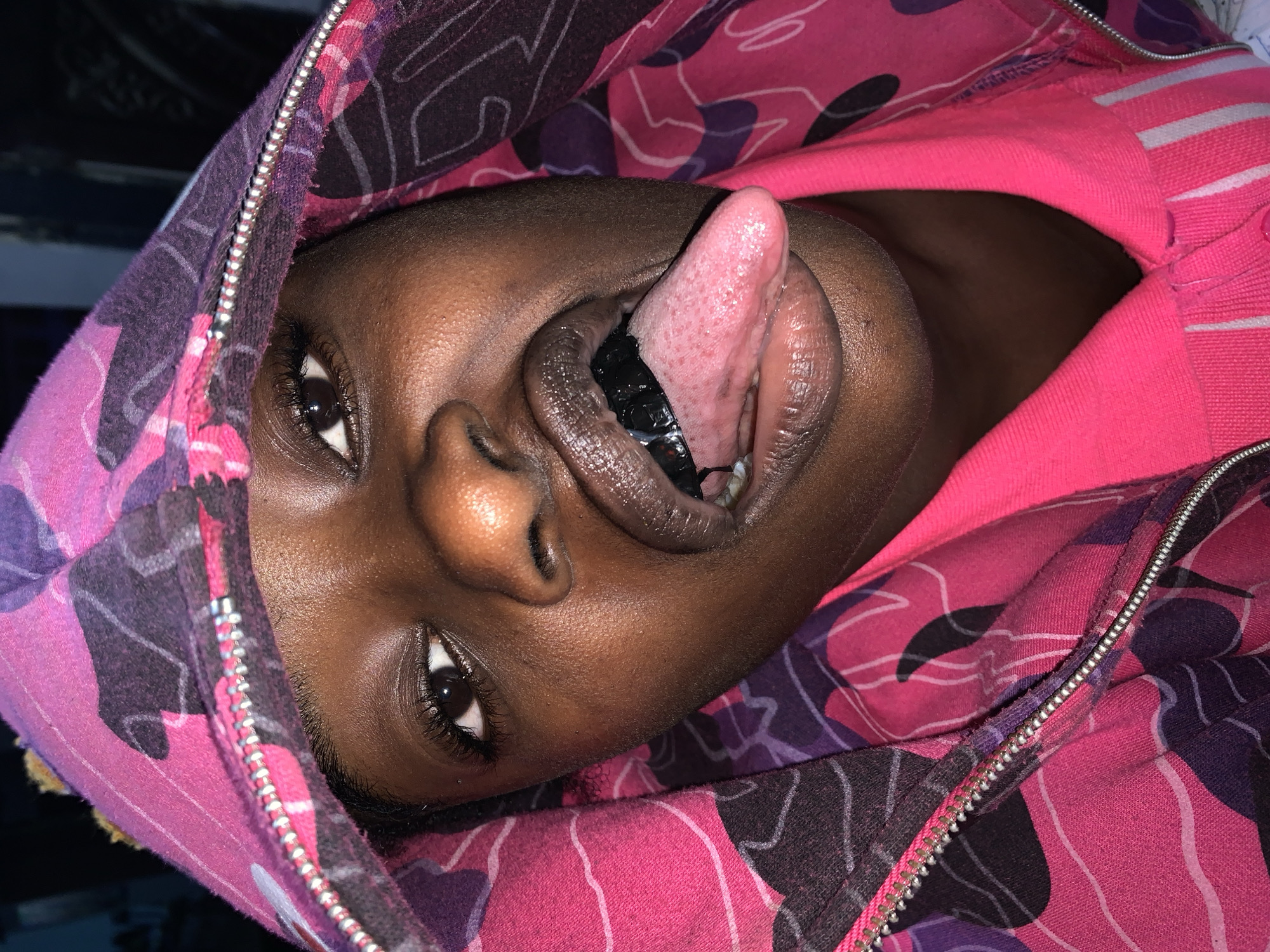 The image shows a close-up, over-the-shoulder view of a young Black person wearing a brightly colored pink and purple hooded jacket. The person is looking directly at the camera, with their mouth open and tongue extended. The tongue is pale pink and appears to have some red spots. Inside the mouth, several dark, rectangular objects are visible, possibly teeth jewelry or decorative items. The person's skin is dark brown and appears to have a slight sheen. The background is dark and indistinct, providing no contextual information about the location. The image was taken in New Stadium, Nigeria.