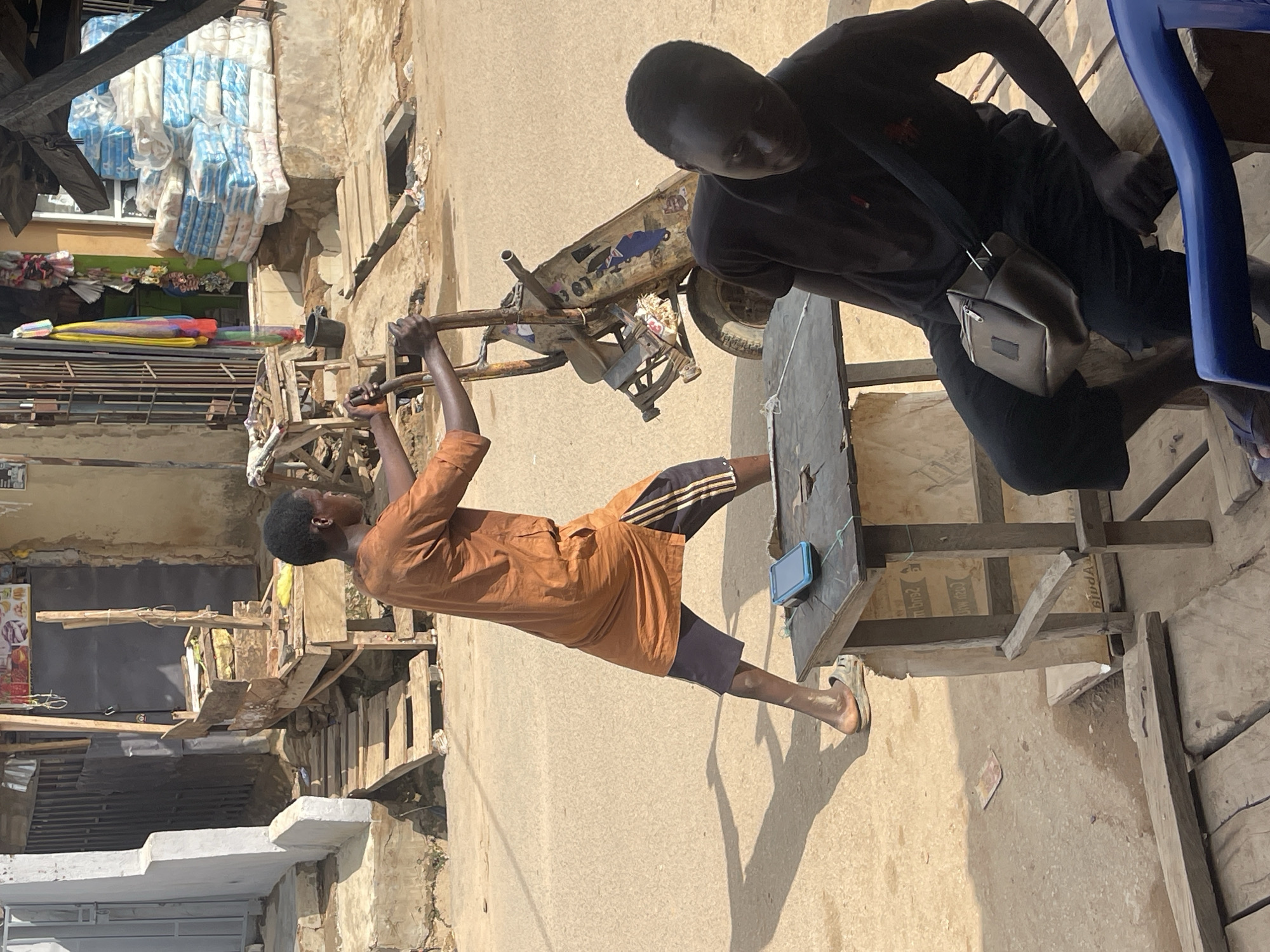 Two men are present. One man pulls a rusted hand truck. He wears a brown shirt and dark shorts with light stripes. The second man stands near a makeshift wooden table. He wears a dark shirt and shorts with a shoulder bag. A blue cell phone sits on the table. The background shows cluttered urban architecture. Piles of wrapped goods are visible atop a nearby building. The location is Abuja, Nigeria.