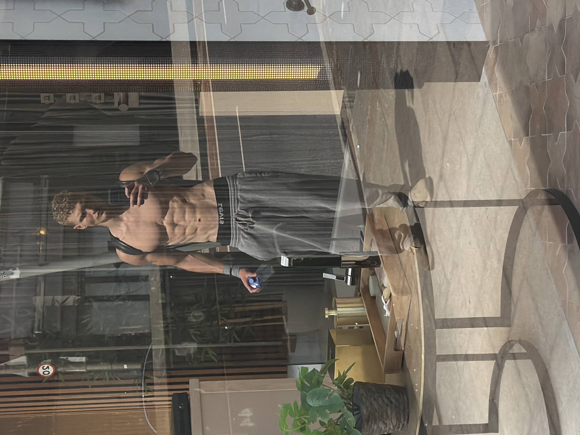 A shirtless man with visible abdominal muscles and blond, curly hair takes a mirror selfie while standing in front of a storefront. He is wearing grey sweatpants and a black backpack. In his left hand, he holds a blue water bottle and what appears to be a black wristband. The man is reflected in a large window, which also reveals the interior of the store, including plants, a golden decorative box, and a wooden tray with small items. The exterior environment shows vertical wooden blinds, a traffic sign with "50" inside a red circle, and a yellow LED light display. The floor outside the store has a patterned tile design. The image is taken from a low angle, capturing the reflection on a table in the foreground. The location is not identifiable as London, United Kingdom.