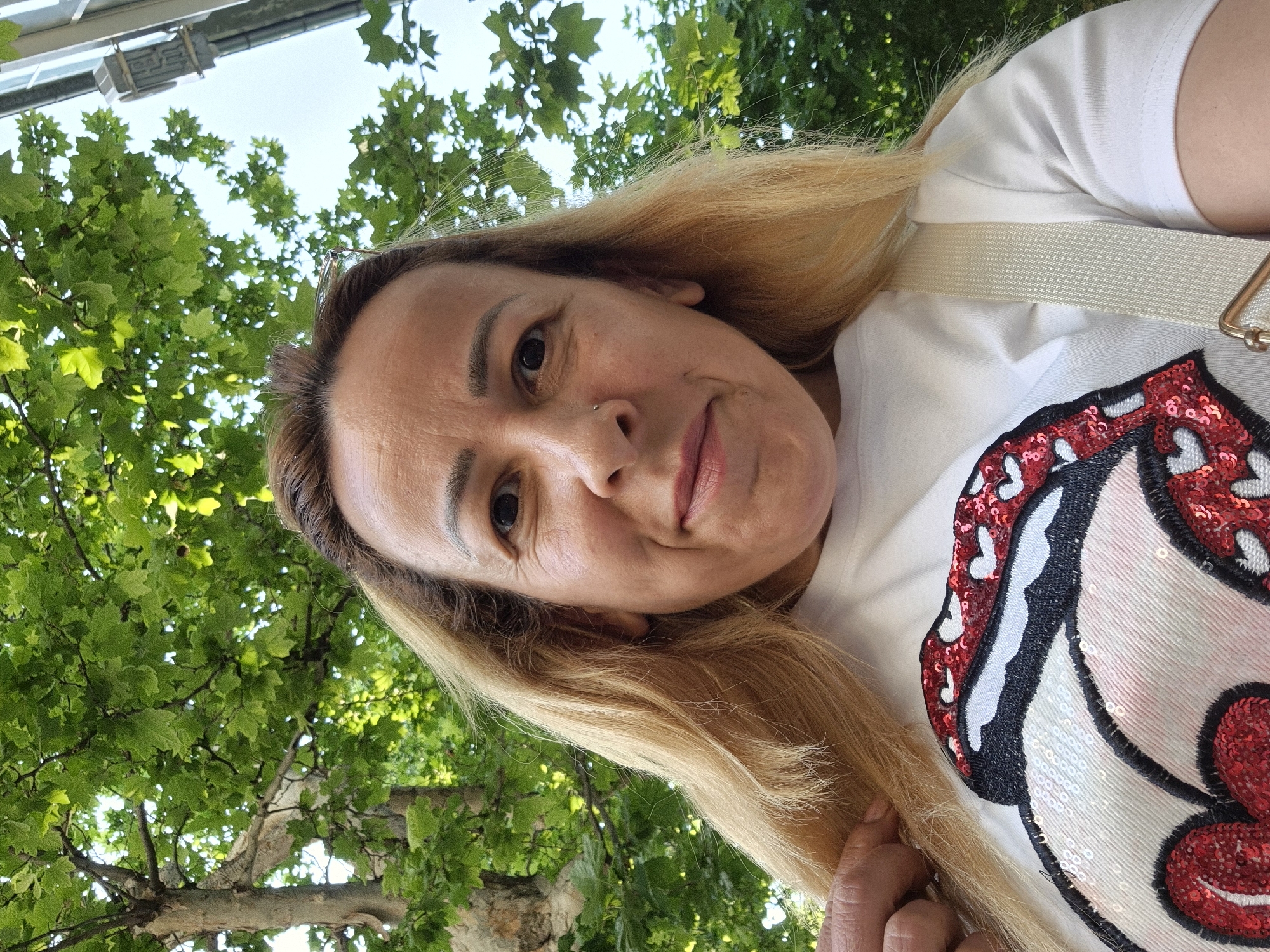 A close-up, low-angle, outdoor selfie shows a woman with blonde hair and fair skin from the chest up. She is looking directly at the camera with a slight smile. Her hair is partially obscuring her face and shoulders. She is wearing a white t-shirt adorned with a large, red and white sequined graphic of lips and hearts. A light-colored strap of a bag or purse is visible across her chest. Behind her, lush green leaves of a tree fill the upper portion of the frame, with a sliver of a building visible on the right. The lighting suggests it is daytime.