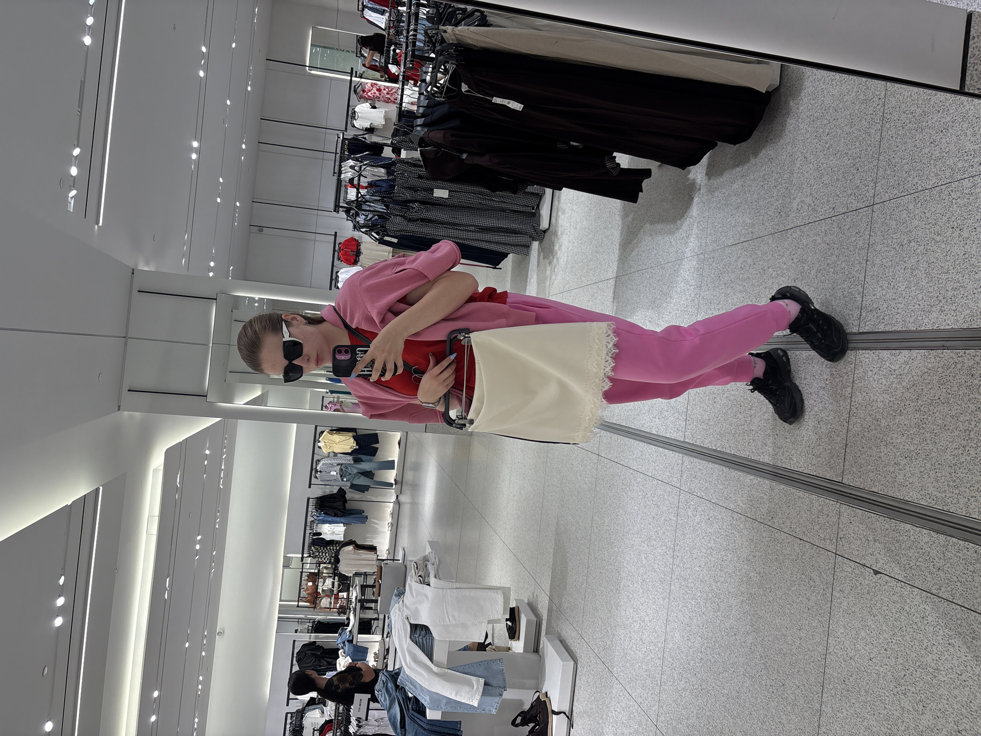 A person is taking a mirror selfie in a retail clothing store. The individual wears sunglasses, a pink outfit, and black shoes. They hold a red purse and a white tote bag. The store has racks of clothing displayed behind the person, and the floor is tiled with a light gray pattern. The location is in Plovdiv, Bulgaria.