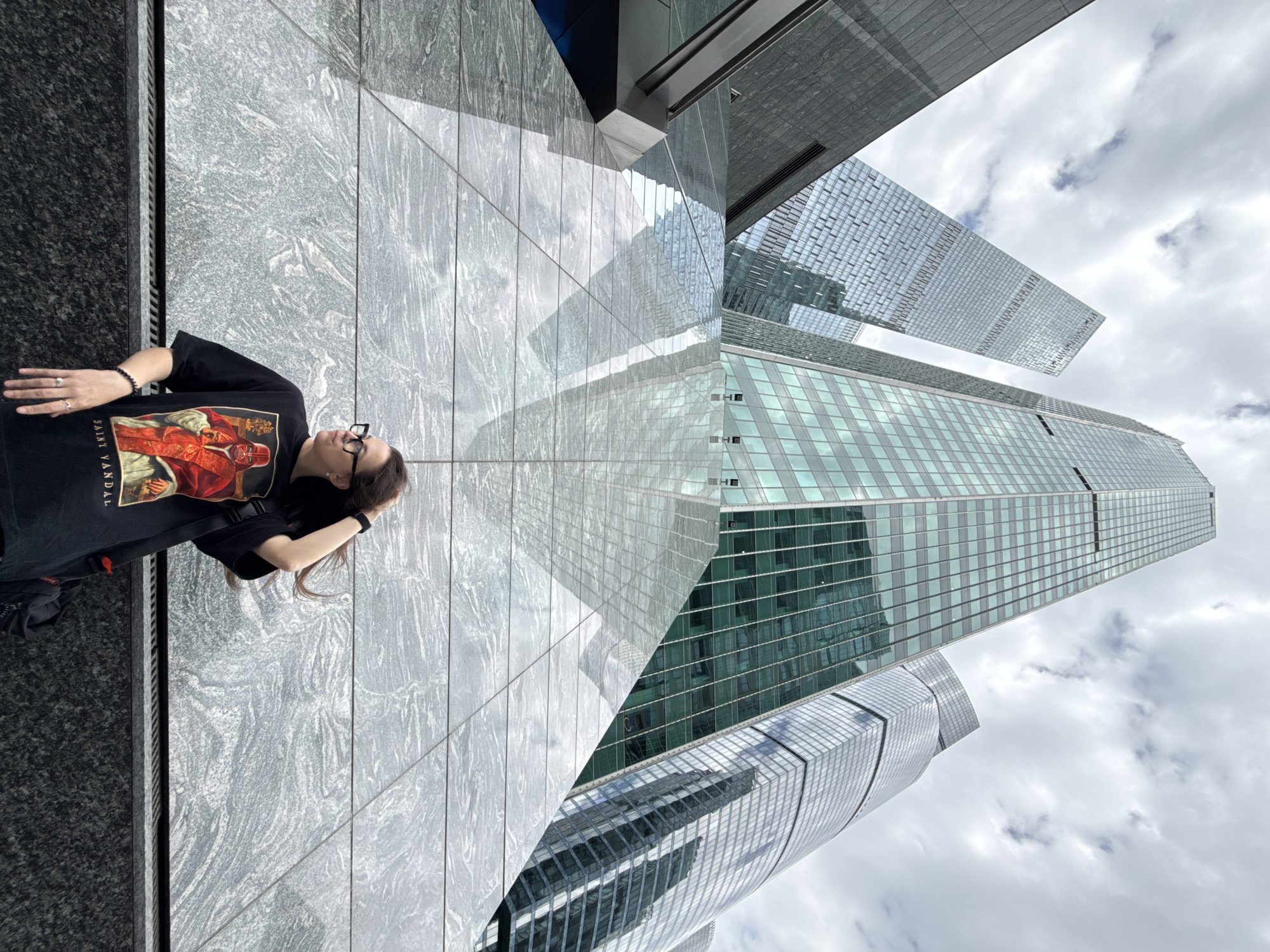 A person wearing glasses and a black t-shirt with a graphic design is lying on a gray marble surface. Behind them are several modern skyscrapers with glass facades, partially reflecting the cloudy sky. The location is Khimki, Russia, based on the mention of it. The buildings appear to be high-rise office or residential structures. The person is interacting with the surrounding environment through their pose, seemingly taking in the architectural scene.