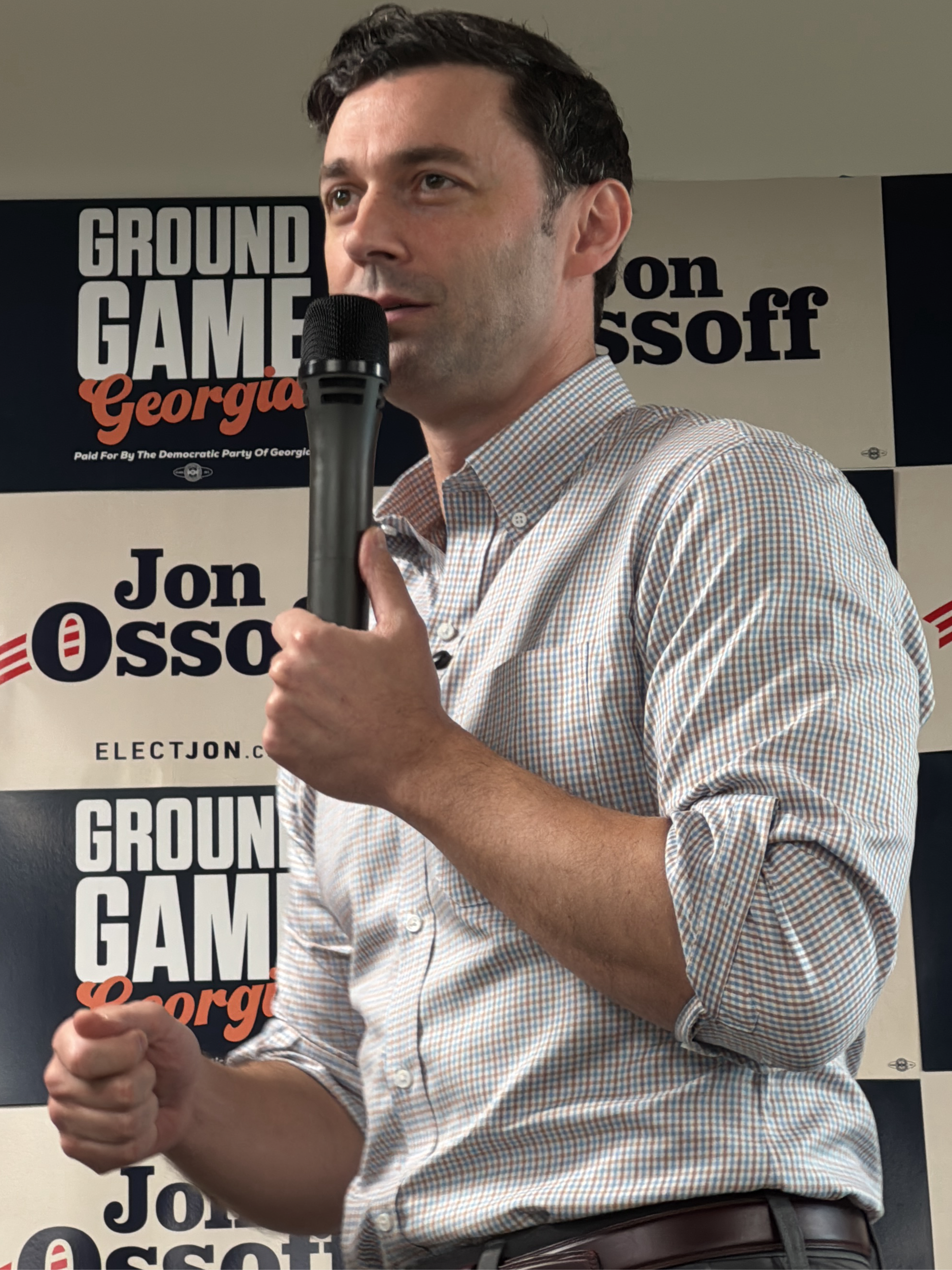 The image shows Jon Ossoff, a United States Senator from Georgia, speaking at an event. He's holding a microphone in his right hand, with his left hand clenched in a fist. He is wearing a checkered button-down shirt. The background features campaign signage with the words "GROUND GAME Georgia", "Jon Ossoff", and "ELECTJON.C". There is also a statement that reads "Paid For By The Democratic Party Of Georgia". Based on the image content of campaign material, it can be inferred that this took place in Decatur, Georgia, United States.