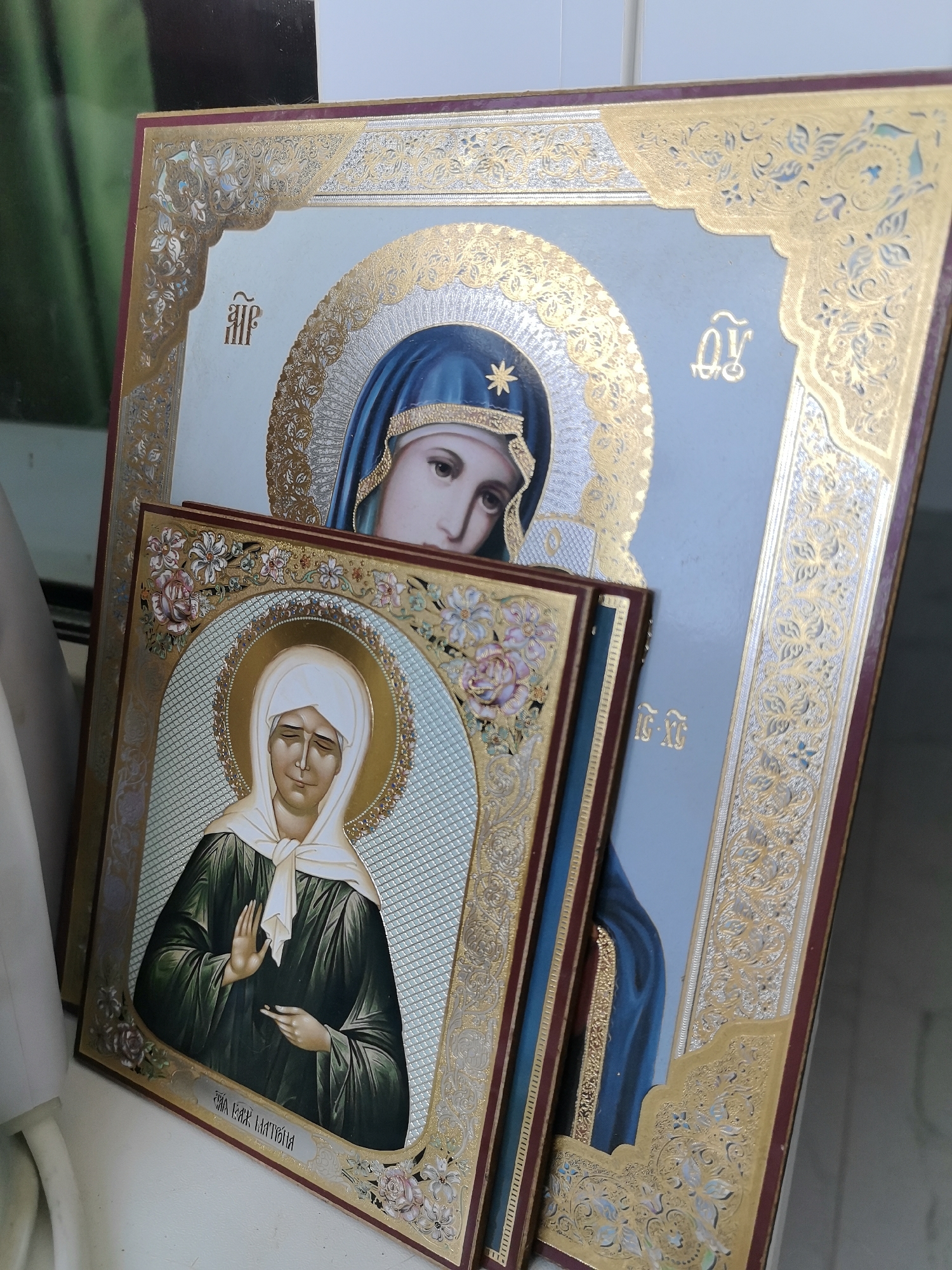 Two religious icons are stacked on a white surface, with a blurred green object in the background. The top icon depicts the Virgin Mary in a blue veil and robe, with a halo and ornate gold trim. The bottom icon features a woman with a white headscarf and green robe, also with a halo and decorative gold framing. Cyrillic script is visible on both icons. The scene appears to be indoors, possibly in a home or a place of worship, in Novoaltaysk, Russia.