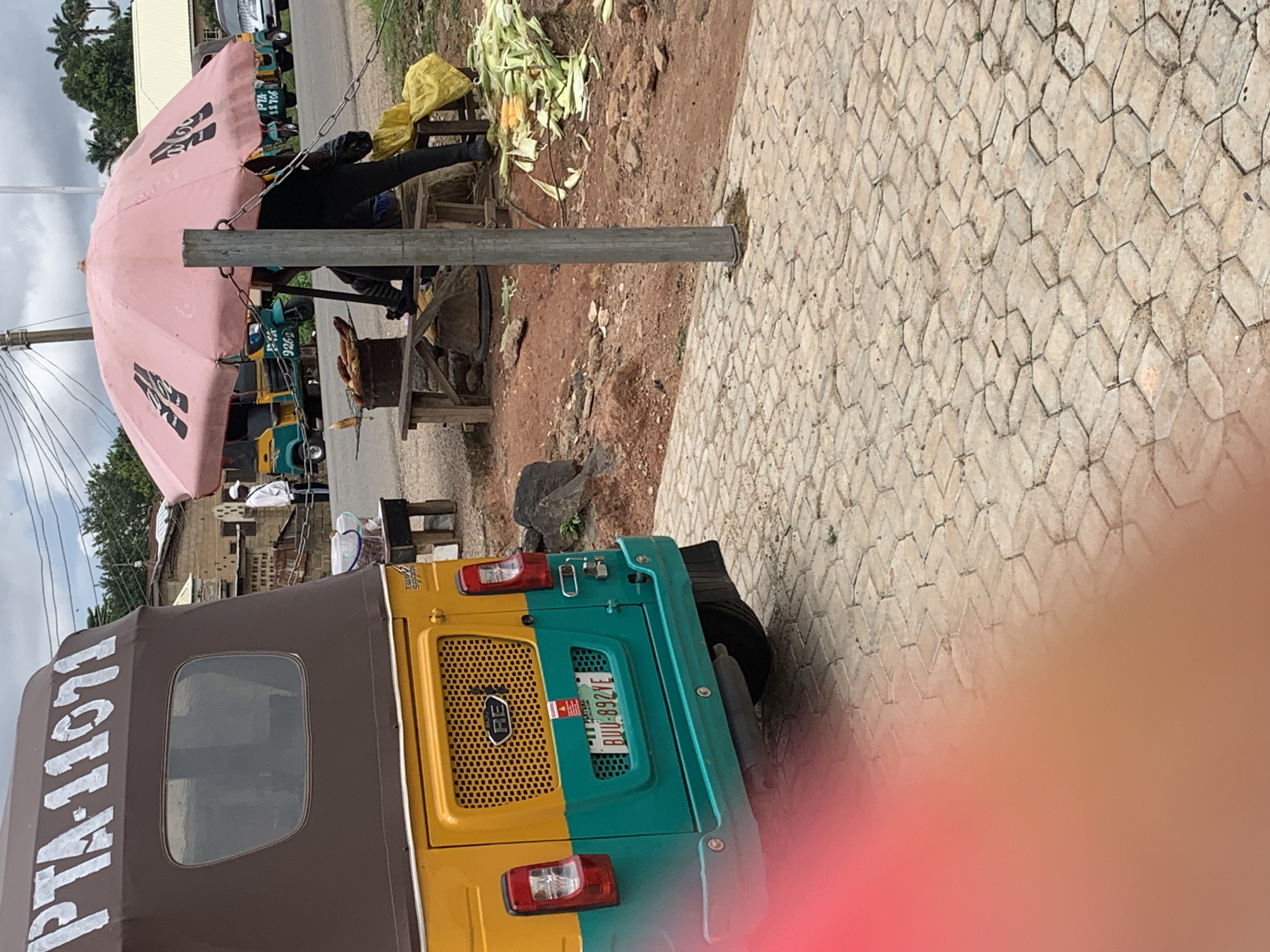 The image shows a scene at what appears to be a roadside market in an urban area. In the foreground, a yellow and teal colored auto-rickshaw is parked, with the license plate reading "BUU-892YE". The vehicle has "PTA-11071" written on its brown canvas roof. Behind the auto-rickshaw, a pink umbrella is set up, advertising "Adidas" with its iconic three stripes logo. Underneath the umbrella, a person dressed in black is seated. In front of them, corn is being roasted over a fire in a metal drum, and a pile of husked corn is visible to the right. Other auto-rickshaws are parked in the background, suggesting a transportation hub or a busy street. A person in a white shirt and a white cap is standing near a building in the mid-ground. The ground is paved with interlocking hexagonal stones. The sky is cloudy.