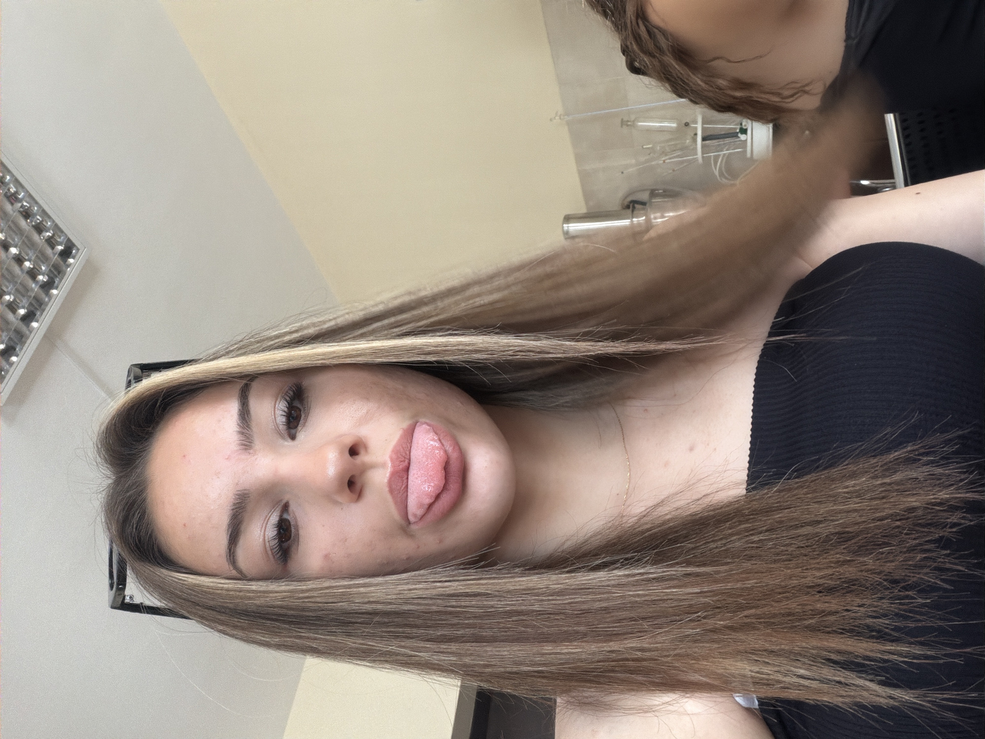 A young woman with long, blonde-streaked brown hair sticks her tongue out at the camera. She is wearing a black off-the-shoulder top. She has a thin gold necklace on. Her face has some acne. In the background, there is a fluorescent light fixture on the ceiling, light yellow walls, a partial view of a metallic appliance, and some lab equipment on a shelf. To the right of the main subject, another person with curly brown hair is visible. The image appears to have been taken indoors. There is no visual information to determine a specific location within the city of Shumen, Bulgaria.