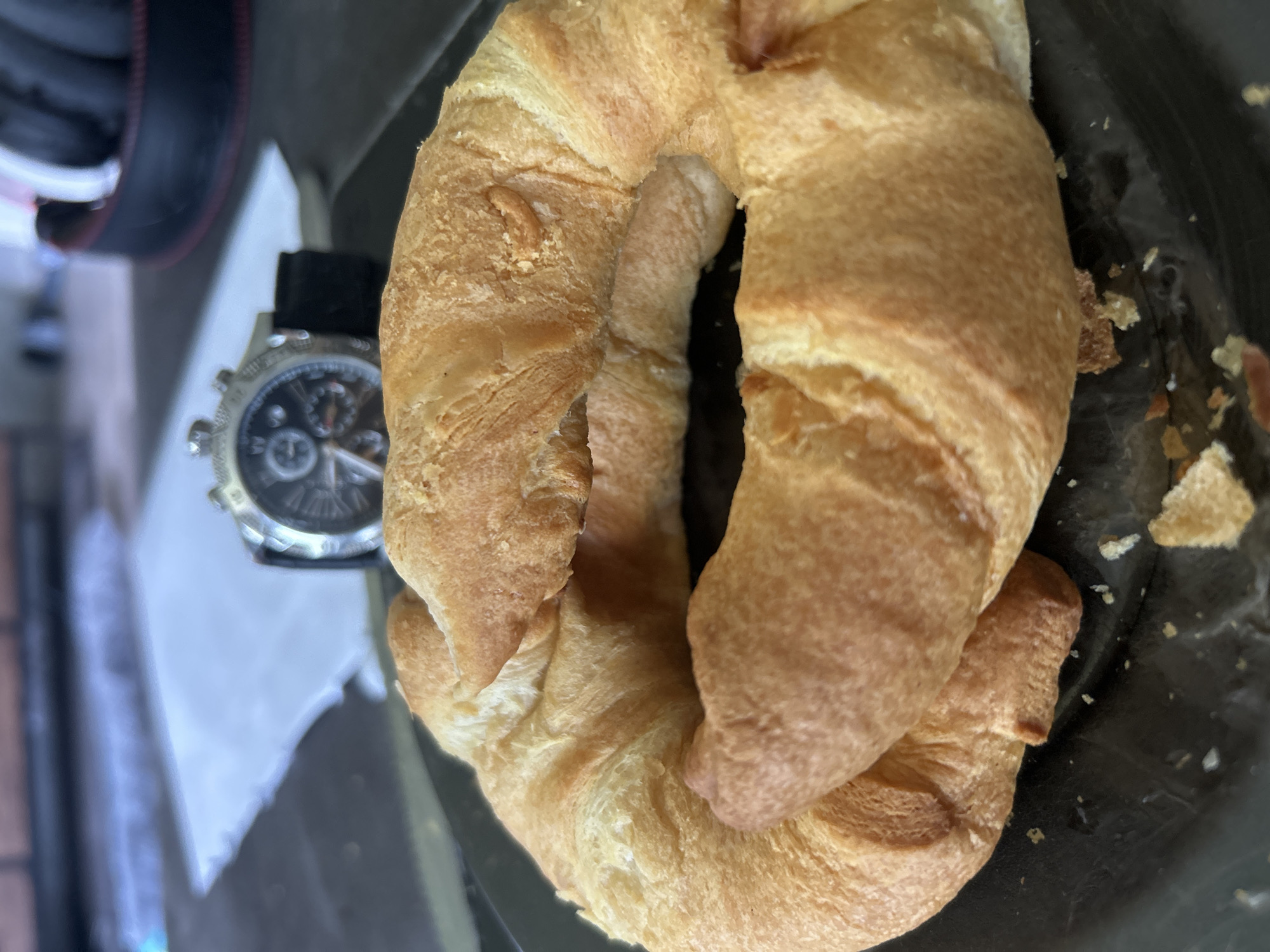 A close-up, eye-level shot captures two golden-brown croissants resting on a dark, possibly black, plate. The croissants are slightly overlapped, with the flaky layers of dough clearly visible. In the background, slightly out of focus, a wristwatch with a dark band and a complex face is positioned on a dark surface. Behind the watch, a white paper object is visible, and further back, a dark, textured surface appears to be a table or countertop. The lighting suggests an indoor or shaded outdoor setting, with a hint of brickwork visible in the far background. The image is devoid of any human presence. The scene is consistent with a breakfast or snack setting, likely in a cafe or home environment in Leiden, Netherlands.