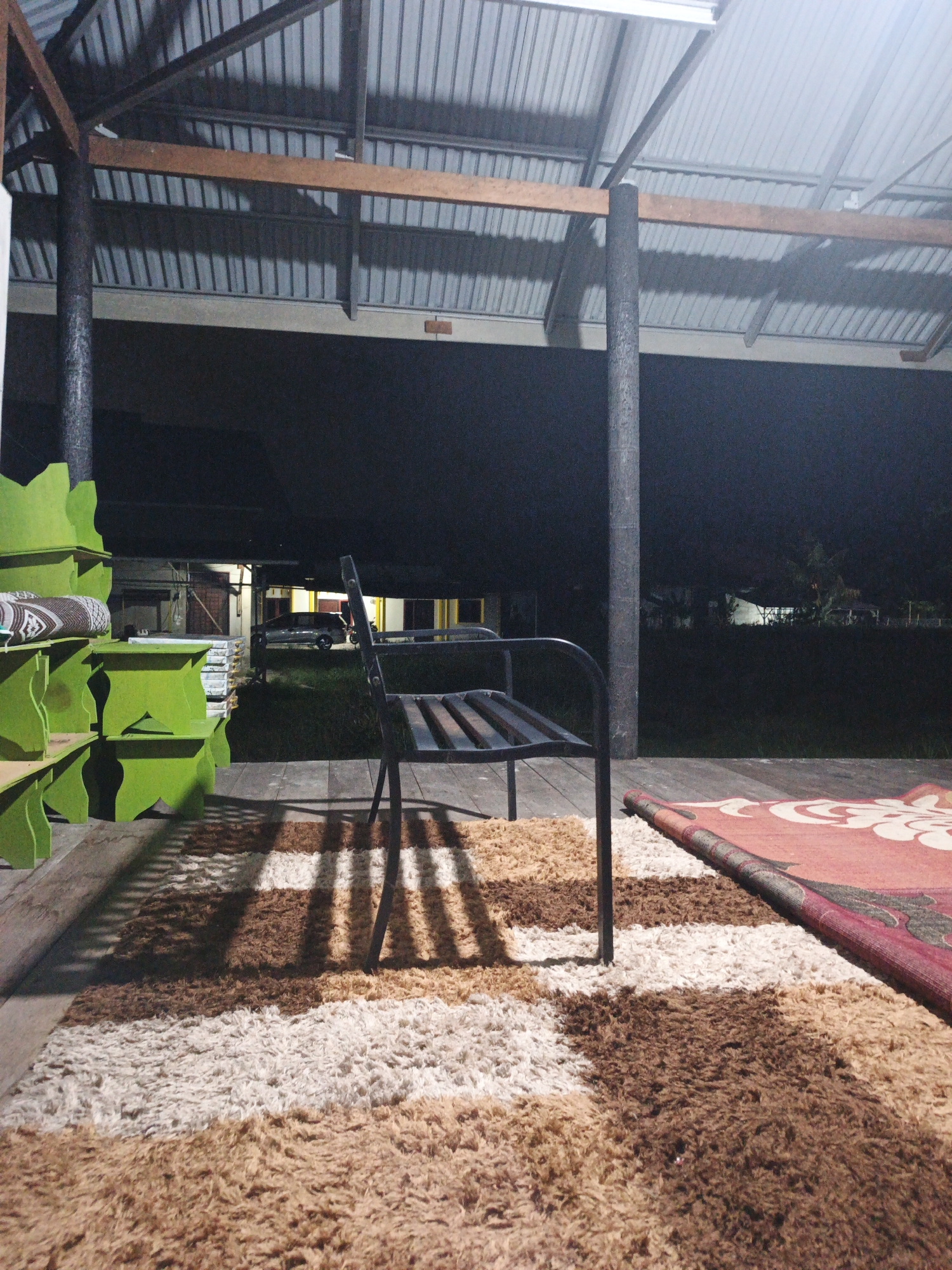 The image depicts a porch area at dusk or night, possibly in Kelapapati, Indonesia, illuminated by artificial light. A corrugated metal roof supported by wooden beams and a thick, dark pole is visible overhead. In the foreground, a wooden deck is covered by a striped shag rug featuring bands of brown, beige, and white. To the right, a portion of a red rug with a floral pattern is partially rolled up. A dark metal bench with a slatted seat sits on the deck, casting shadows from the roof onto the rug. To the left, bright green, open-shelving units are stacked, with a rolled-up mat or fabric visible on the top shelf. In the background, a dark curtain or wall obscures the view, but glimpses of buildings with lights and a parked car suggest a residential or commercial area. There are no people present in the image.
