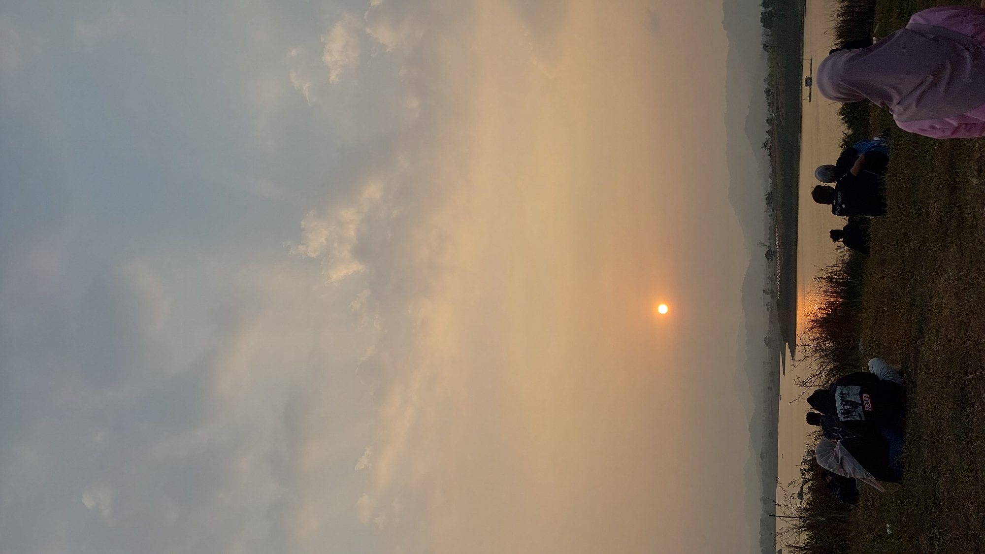 A vertical, eye-level shot captures a tranquil sunset scene by a body of water. The sun, a small, bright orange orb, is positioned in the lower-center of the frame, casting a warm glow across the hazy sky and reflecting on the water. Silhouetted against the sunset are several individuals and groups of people sitting on a grassy embankment overlooking the water. The landscape features rolling hills in the distance, partially obscured by the haze. The foreground is dominated by dry grass and the figures of the seated people. In the lower-right corner, a person wearing a pink hijab and a light-colored top is partially visible, facing away from the camera. To the left, a group of people are huddled together, with one individual wearing a black hoodie with a graphic on the back. The overall atmosphere is one of peaceful observation of the natural spectacle.