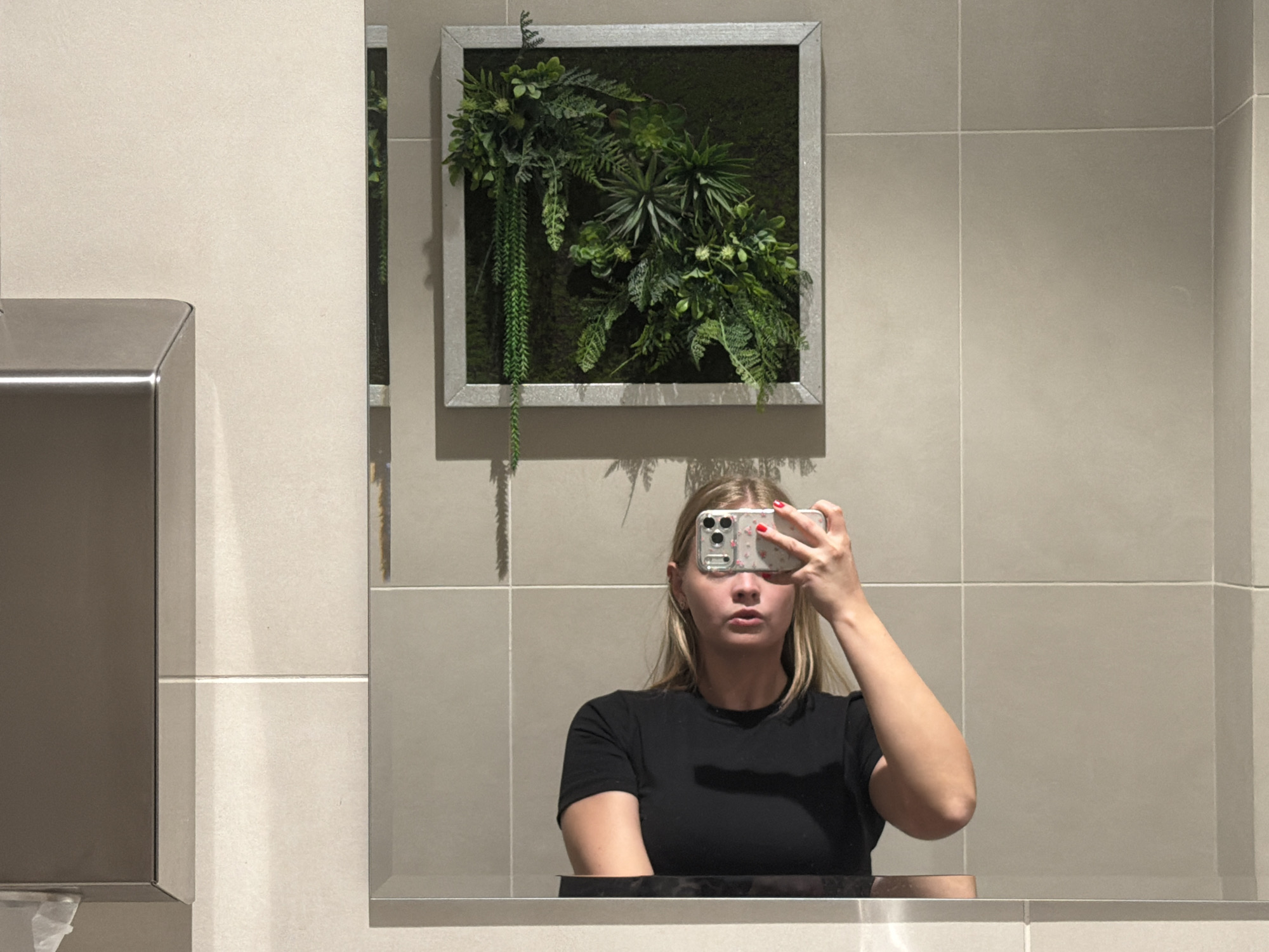 A woman with long blonde hair wearing a black t-shirt is taking a selfie in a bathroom mirror. The reflection shows a framed square moss art piece hanging on the wall behind her. The bathroom walls are covered in large, light-colored tiles. A metallic paper towel dispenser is visible on the left side of the mirror's reflection. The location is unknown, and Izhevsk, Russia is not confirmed.