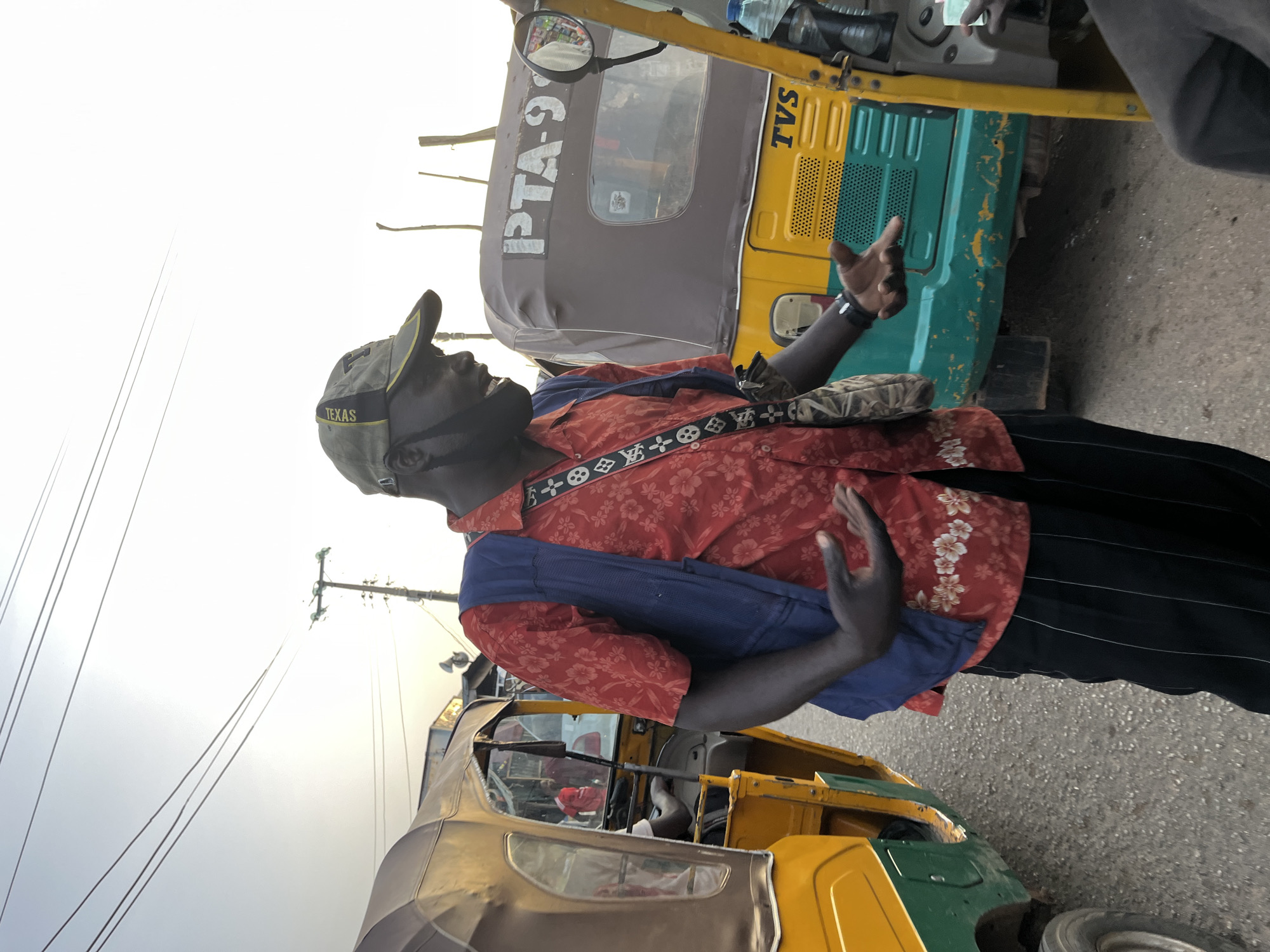 In Gura Top, Nigeria, a Black man wearing a red floral shirt, a dark vest, black striped pants, and a baseball cap is standing by a yellow and green auto-rickshaw. He is gesturing with his left hand and speaking. Another auto-rickshaw, with the license plate "PTA-9", is visible behind him. The scene appears to be a street or roadside in Nigeria.