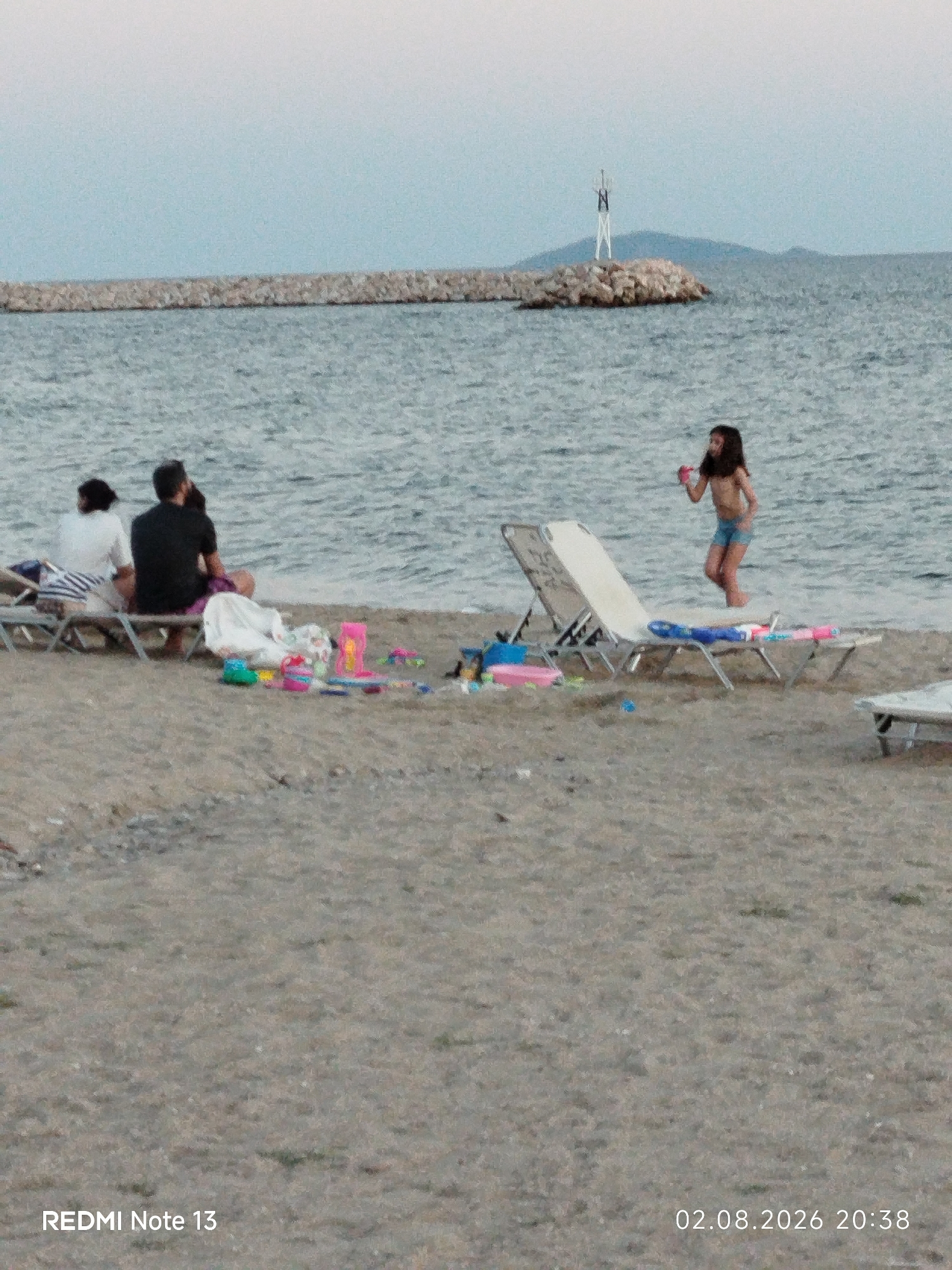 The image depicts a beach scene in Sithonia Municipal Unit, Greece. In the foreground, the sandy beach is visible with scattered beach toys and two lounge chairs. In the middle ground, two individuals are seated on lounge chairs facing the water, with another person in dark clothing behind them. To the right, a young girl in a swimsuit is standing in the shallow water, holding a pink object. In the background, the sea stretches out to a horizon with a breakwater made of rocks and a small navigational marker on a rocky outcrop. The sky is overcast and a pale blue. The timestamp "02.08.2026 20:38" and "REDMI Note 13" are overlaid on the bottom of the image.