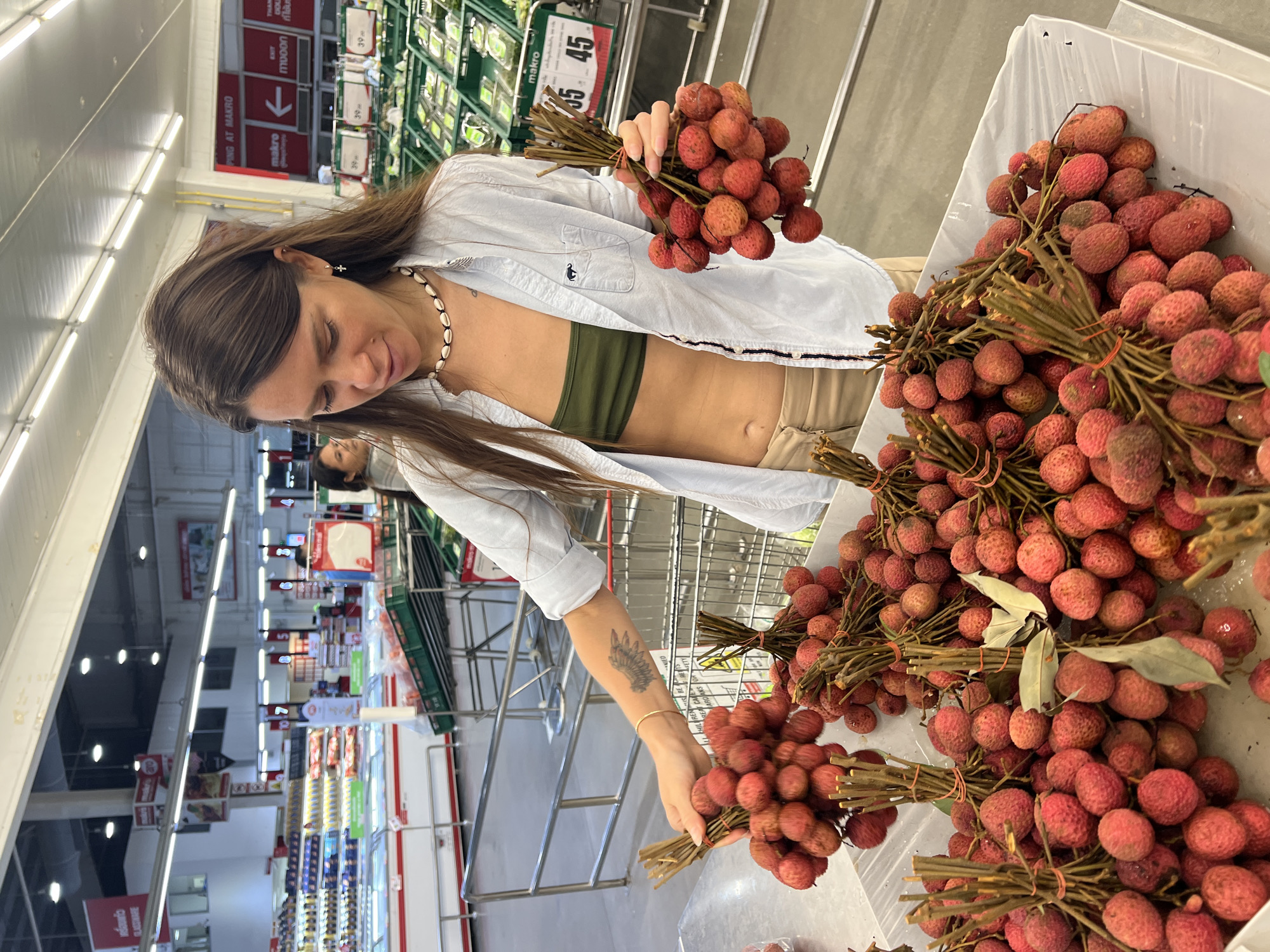 A woman in a light-colored button-down shirt and khaki shorts is shown in a supermarket setting, possibly located in Baan Bo Phut, Thailand. She stands beside a table laden with bunches of lychees, examining the fruit. She is holding one bunch in her left hand and is reaching for another with her right hand. Her dark hair is parted down the middle, and she wears a shell necklace. An olive green tank top can be seen beneath her shirt. A tattoo is visible on her left forearm. The scene includes the interior of a large retail space, with shelving stocked with various items and price signage in the background, specifically a sign with the number "45". There are shopping carts and other shoppers visible. The overall setting indicates a busy supermarket environment.