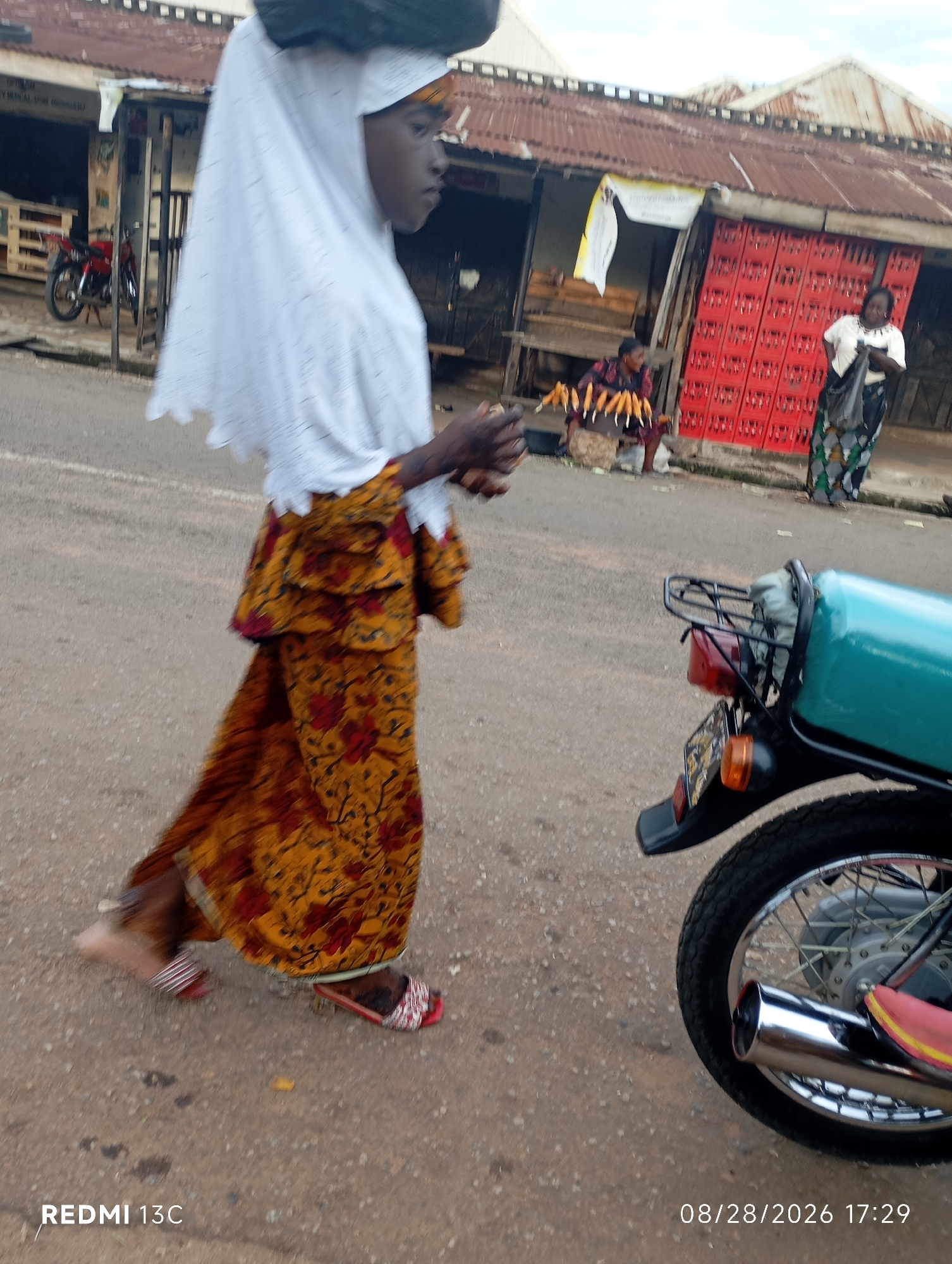 In Shendam, Nigeria, on a dusty street in front of a row of market stalls, a young person walks past a teal-colored motorcycle. The individual wears a white headscarf that obscures their hair, and a long, patterned skirt in shades of yellow and red. They are carrying a large, dark object balanced on their head. In the background, two women are visible at one of the stalls. One woman, dressed in black and white, stands holding a black plastic bag, while another woman sits behind a display of corn on the cob. Red crates are stacked near the stalls, and a red motorcycle is parked further down the street. The sky is overcast.