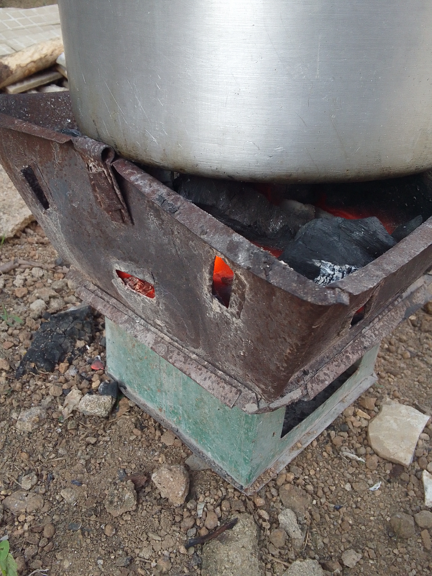 A close-up shot captures a cooking scene outdoors. The focal point is a large, metallic pot placed atop a rusted, rectangular metal brazier. The brazier, with its weathered texture and visible signs of wear, is filled with glowing charcoal embers, indicating an active fire. The heat from the coals is visible through the openings on the sides of the brazier, casting a warm orange hue.
The brazier itself is supported by a green, box-like structure, possibly a stand or a base, also showing some signs of age. The surrounding ground is rough and uneven, covered with a mix of soil, small stones, and scattered pieces of burnt wood or charcoal. In the background, a few logs or pieces of wood are visible, suggesting a rustic or rural setting. A hint of a tiled surface can be seen in the upper left corner.
The image appears to be taken during daylight, though no specific time of day can be definitively determined. The lighting suggests an outdoor environment, possibly a kitchen area or a cooking spot in a less developed area. There are no people visible in the frame, and the focus is entirely on the cooking apparatus and the immediate surroundings. The overall impression is one of a functional, perhaps traditional, method of cooking. No text is visible in the image.