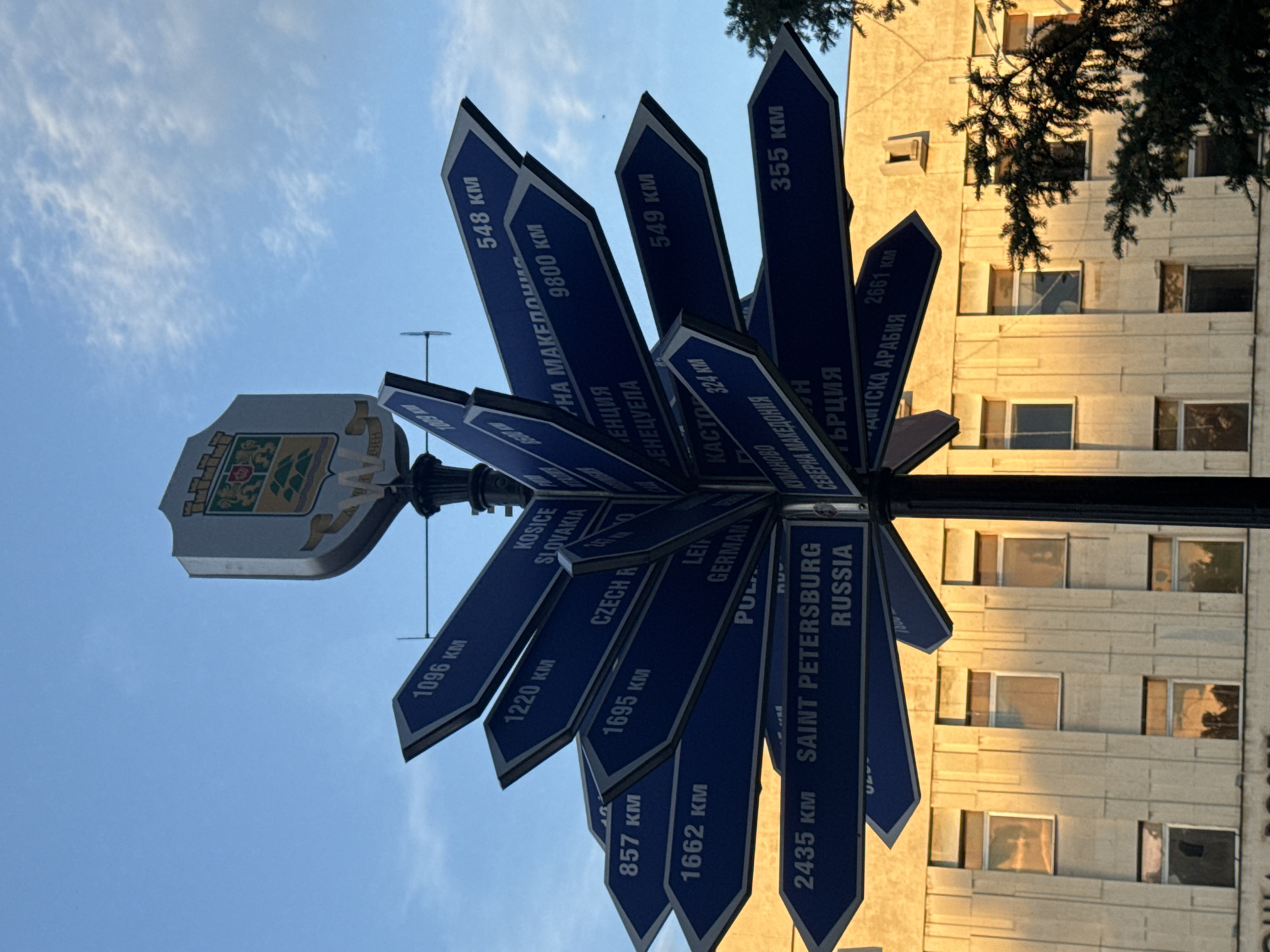 The image shows a directional signpost in Plovdiv, Bulgaria. The signpost features multiple blue signs pointing in different directions, each indicating a city and the distance to it in kilometers. Some visible cities include Kosice (Slovakia), Saint Petersburg (Russia), Kastoria (Greece), and several locations within North Macedonia. Above the signs is the coat of arms of Plovdiv. The background shows a building with numerous windows and a tree with visible branches. The sky is blue with some clouds, suggesting it is daytime.