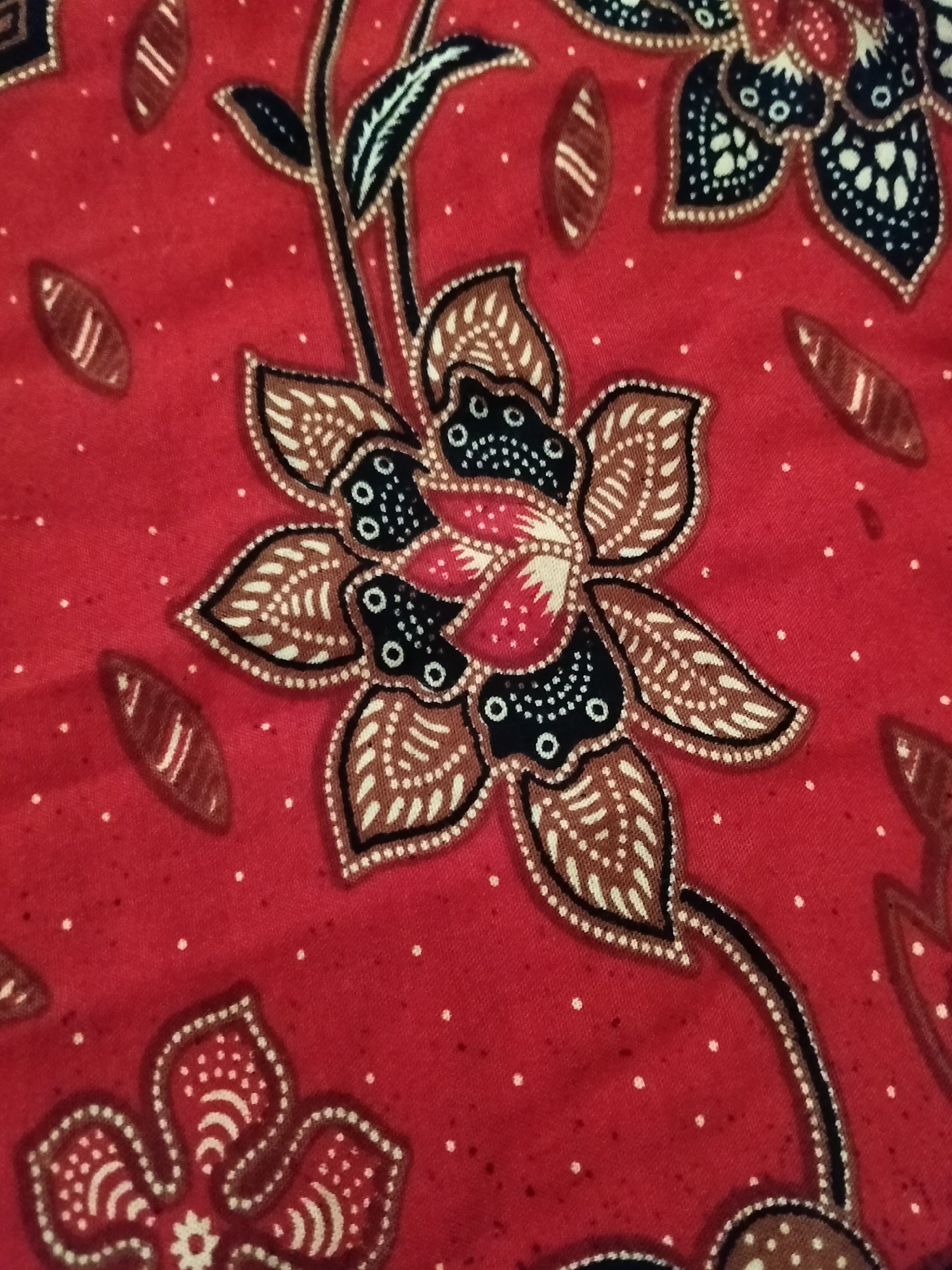 The image is a close-up, overhead shot of a red batik fabric with a floral motif. The fabric features intricate patterns in shades of brown, black, and white, with small white dots scattered across the red background. The design includes stylized flowers with multiple petals and leaves, connected by thin, dark lines. The texture of the fabric is visible, with slight wrinkles and variations in the dye. The scene is devoid of people, actions, or interactions, as it focuses solely on the textile. The location in Gempolan, Indonesia, is noted as the origin of the fabric.