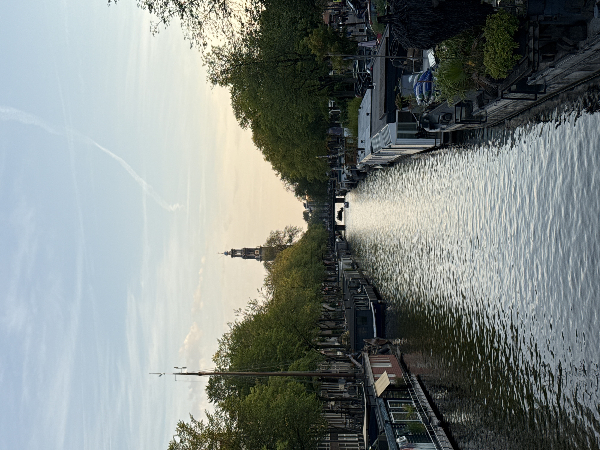 The image depicts a canal in Amsterdam, Netherlands, with houseboats lining both sides. The water is calm and reflects the overcast sky. Trees with lush green foliage grow along the canal banks, creating a shaded atmosphere. In the distance, a tall church tower with a clock face is visible, a common architectural feature in Amsterdam. A few boats are seen on the canal, and parked cars and bicycles are visible on the street behind the trees on the right. The scene appears to be captured during the late afternoon or early evening, given the soft, diffused light and the gentle glow in the sky. The overall impression is one of tranquility and urban charm, showcasing a typical Amsterdam canal scene.