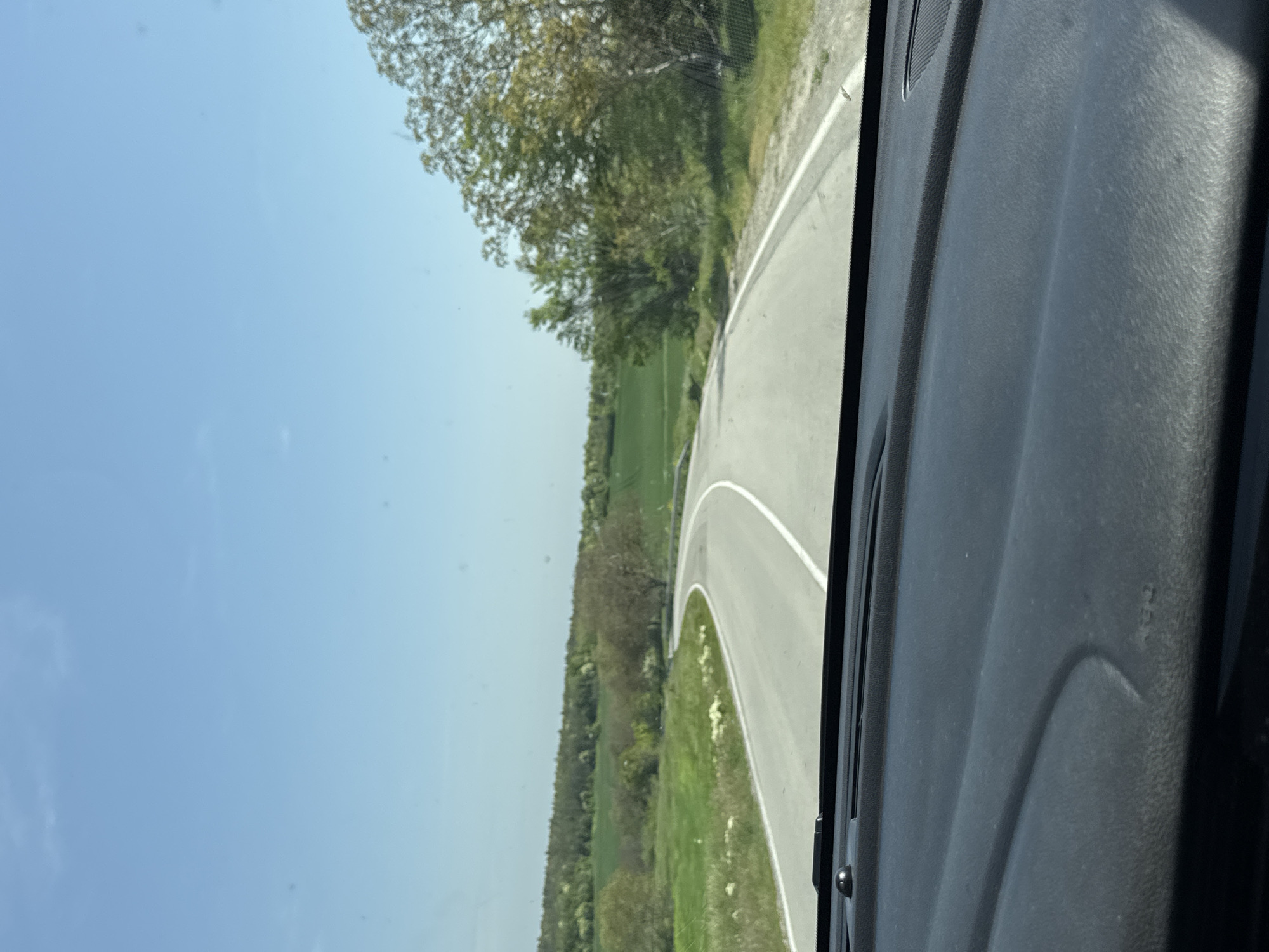 The image shows a view from inside a vehicle, likely a car, as it navigates a curved road. The dashboard of the vehicle is visible in the foreground. Beyond the windshield, the road winds through a rural landscape with greenery and trees bordering the roadside. The sky is blue and clear. The exact location in Royak, Bulgaria is not visually determinable. There are no people visible in the scene.