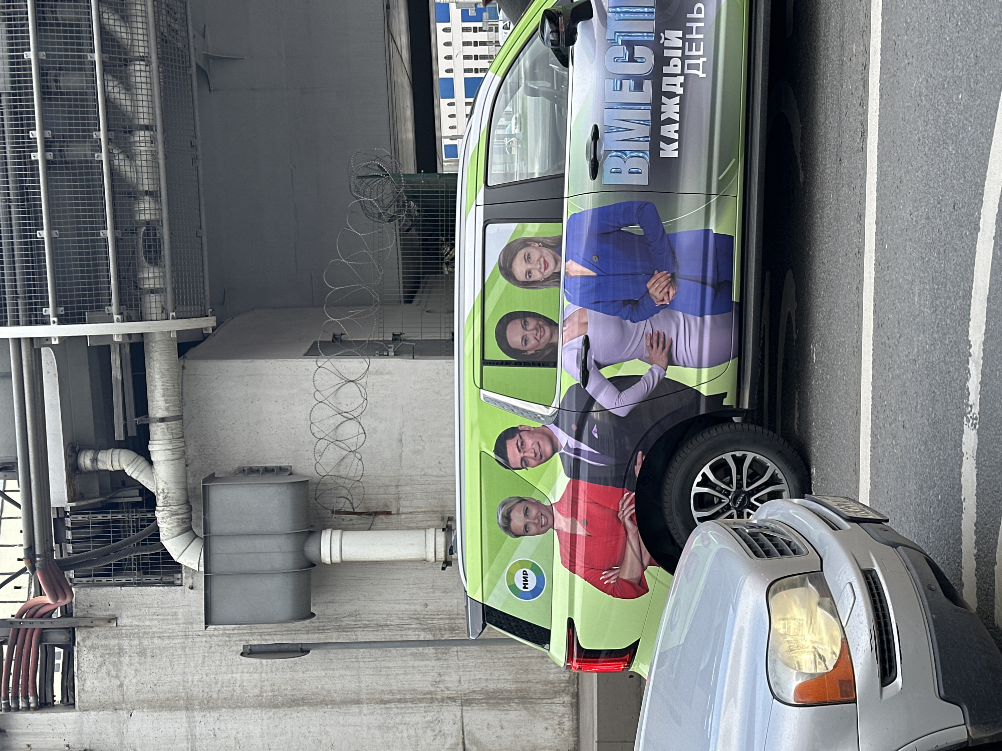 The image captures a parked van alongside a gray car, set against an industrial backdrop in what appears to be a city underpass or a space defined by concrete structures. The van's exterior is distinctive, adorned with a green-themed political advertisement featuring four individuals and the Russian slogan "ВМЕСТЕ КАЖДЫЙ ДЕНЬ," which translates to "TOGETHER EVERY DAY." A political party emblem is visible at the rear of the van. Above and behind the vehicles, a concrete wall is topped with a metal cage and barbed wire, suggesting a secure or restricted area. Piping and industrial ventilation equipment are mounted on the wall. The presence of road markings indicates the vehicles are parked along a designated street. Without additional context or identifiers, the exact location within Moscow cannot be definitively confirmed, but the combination of industrial architecture and Russian-language advertising points to a probable location within the city.