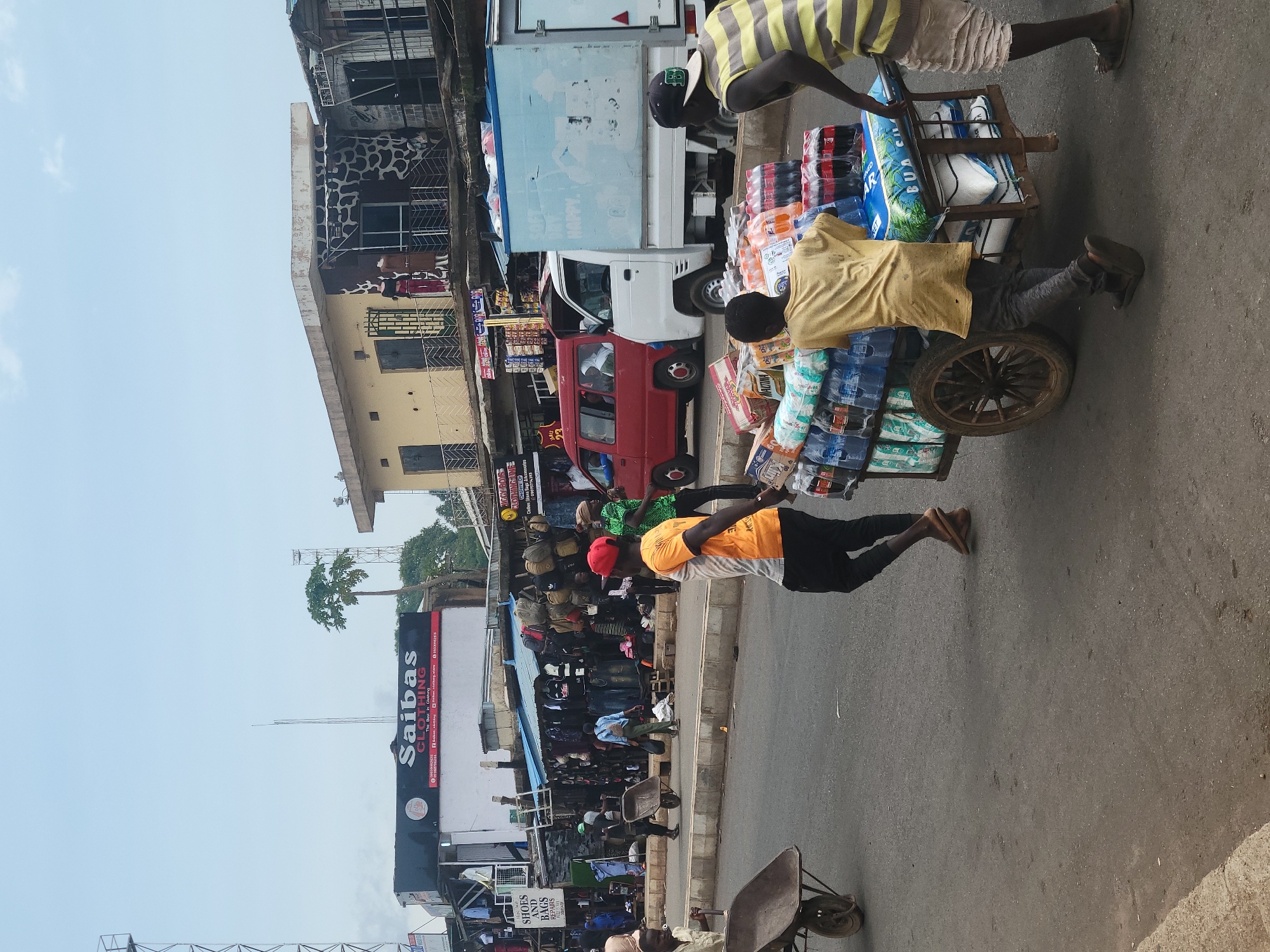The image depicts a street scene in a city, likely a marketplace, with various individuals engaged in activities related to commerce and transport. In the foreground, two young men are pushing a cart loaded with bottled beverages and possibly other goods. One is wearing a yellow t-shirt and dark pants, while the other wears a striped t-shirt and shorts. A wheelbarrow is visible in the lower left quadrant of the image.
Behind the cart, several other people are walking or standing. A man in an orange t-shirt and black pants is walking away from the camera, and another person in a green shirt is also visible. A red van is parked on the street, and a white truck with a blue cargo box is to the right of the van.
The background shows a row of buildings that appear to be shops or stalls. One building has a prominent sign that reads "Saibas CLOTHING". Merchandise, including clothing and bags, is displayed outside some of the establishments. The sky is overcast, and the overall impression is of a busy urban environment with commercial activity.