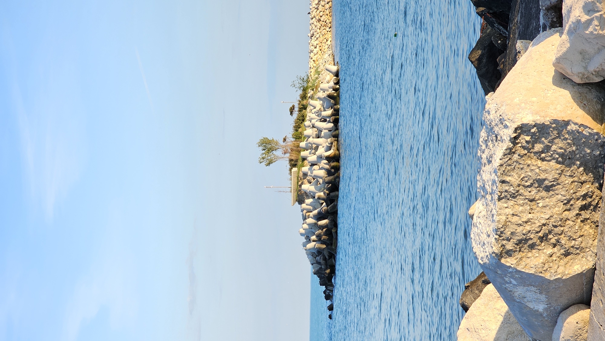 The image depicts a coastal scene in Varna, Bulgaria, specifically a breakwater or pier constructed of large stones and concrete tetrapods. The sea is calm and blue, stretching out towards the horizon where it meets the sky. A small strip of land with sparse trees and vegetation sits on the breakwater. The sky is clear and blue with minimal cloud cover. The photo is taken during daylight, with the sun illuminating the stones and casting shadows.