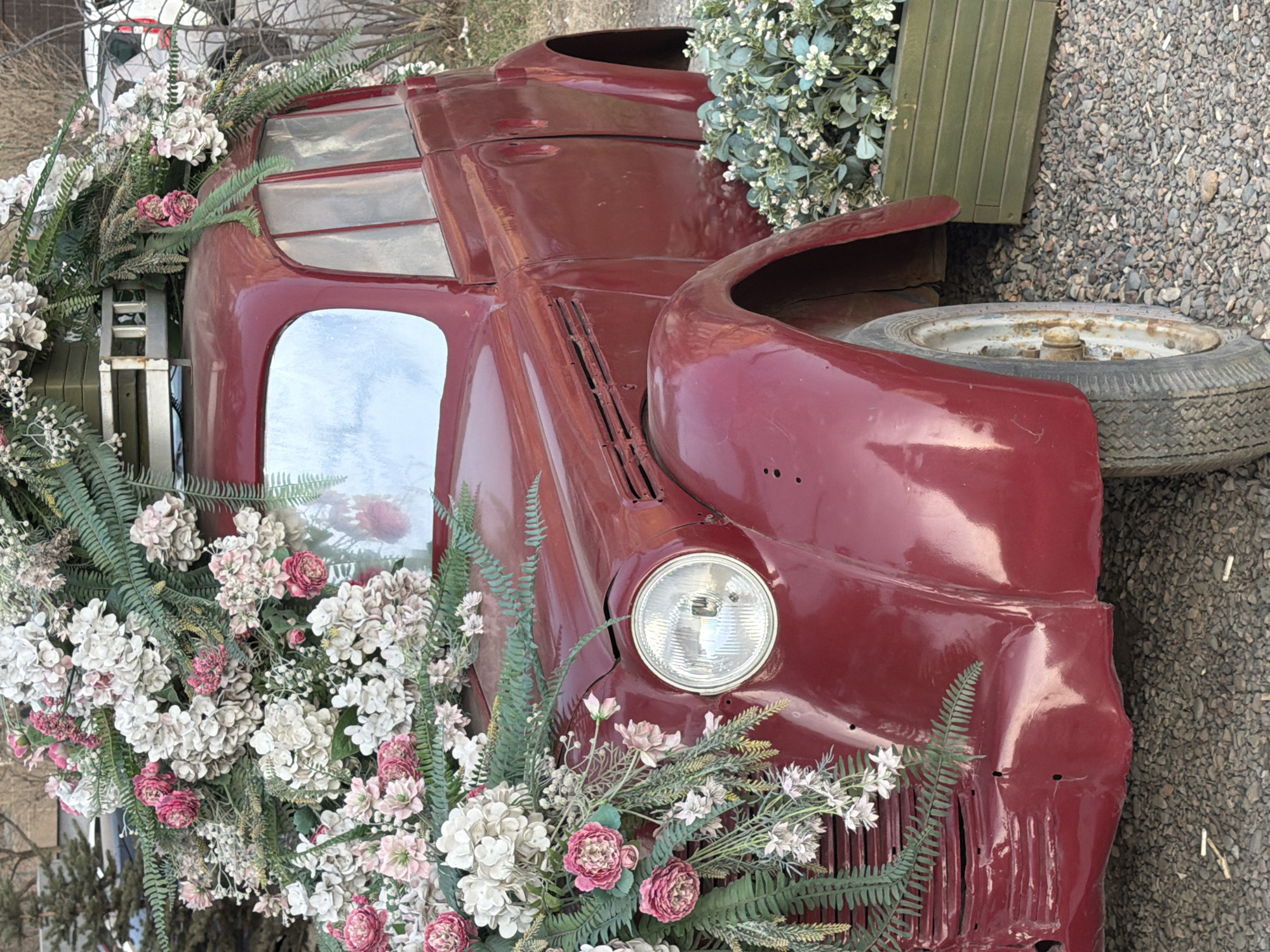 The central subject of the image is a vintage, dark red (maroon) utility vehicle or truck, heavily adorned with artificial white, cream, and pink flowers, along with various green ferns and foliage. The vehicle is oriented with its front towards the bottom-left of the frame, showcasing its circular headlight, part of the grille, the hood, fender, and a side window of the cabin. The car's paint has a glossy finish, though some minor wear and reflections are visible. An exposed, worn spare tire on a rusty wheel hub is positioned near the front passenger side. An olive-green wooden crate rests on the ground behind the vehicle's front section. The ground consists of small, grey gravel. In the blurred background, faint outlines of other structures or objects can be discerned.
There is no active event taking place; the vehicle serves as a static decorative installation or a large-scale planter.
Notable details include the soft, diffused daytime lighting, suggesting an overcast sky or indirect sunlight, as harsh shadows are absent. The extensive floral arrangements create a whimsical and charming contrast with the rustic, aged appearance of the vehicle, implying a creative reuse of the car for aesthetic purposes. The overall ambiance evokes a sense of nostalgic beauty.
There is no visible text in the image.
Location context: Krasnoyarsk, Russia.