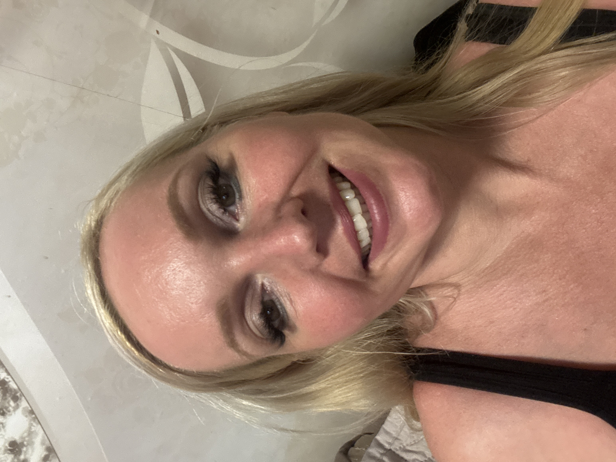 The media file is a close-up, rotated selfie of a fair-skinned woman with blonde hair. She is wearing dark clothing, possibly a tank top or bra. Her makeup includes dark eyeliner and mascara, and her lips are a rosy pink. She appears to be smiling with her teeth showing. The background is out of focus and appears to be a patterned surface, possibly wallpaper or fabric. There are no visible text elements in the image. The lighting suggests an indoor setting, and given the context of Komsomolsk-on-Amur, Russia, it is likely daytime. The overall emotion conveyed is one of happiness or playfulness.