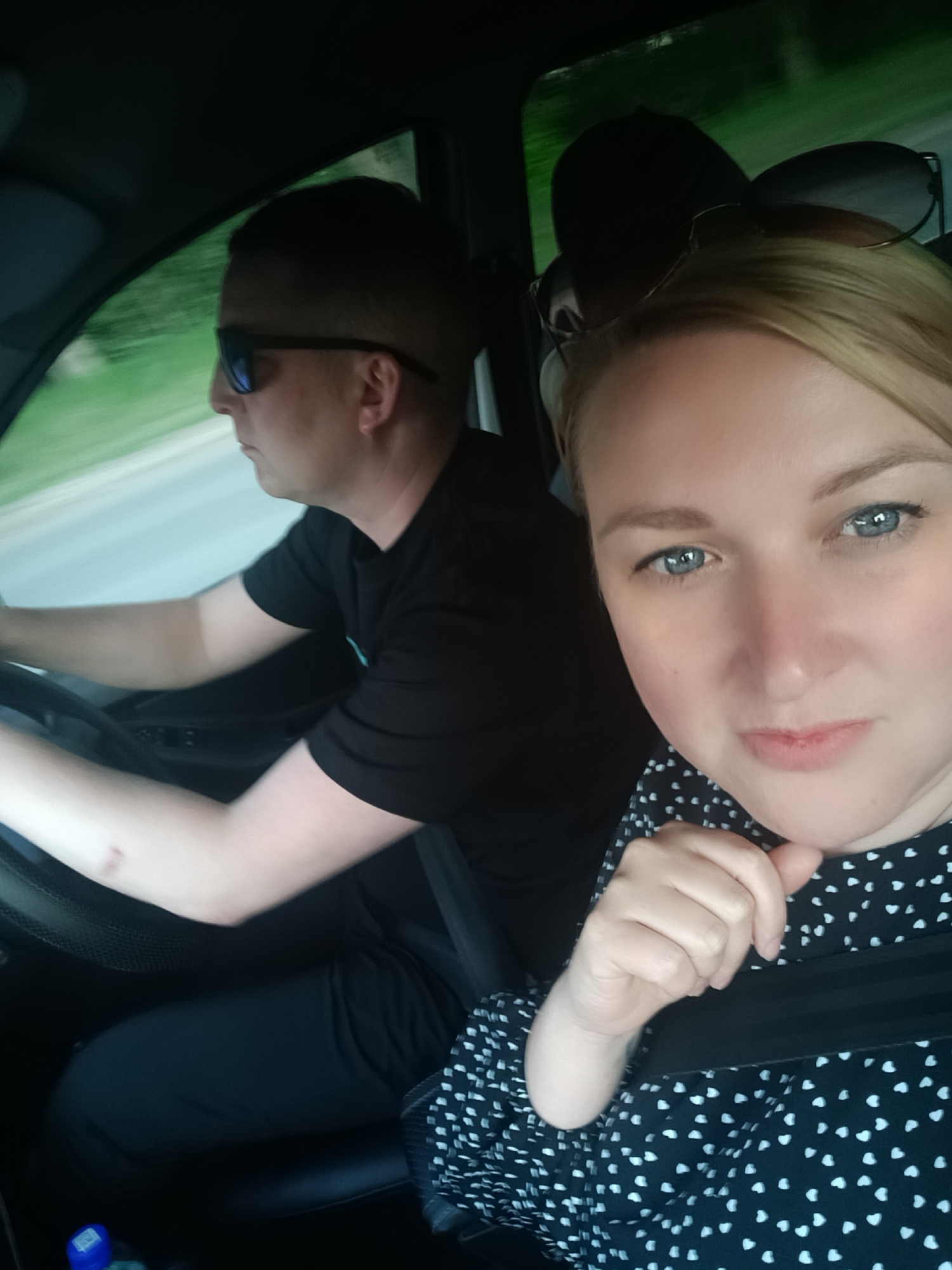 A woman with blonde hair and a patterned black-and-white shirt is taking a selfie in the passenger seat of a car. She is wearing a seatbelt. A man wearing sunglasses and a black t-shirt is driving the car. The car appears to be moving on a road with trees visible outside the window. The location is impossible to identify with just this image.