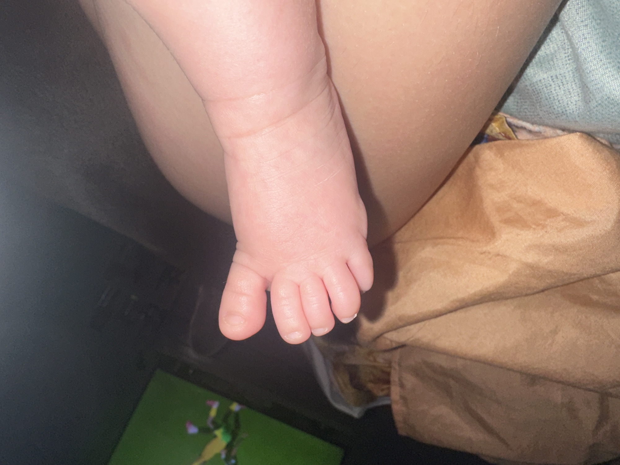 In an indoor environment, a close-up shot focuses on the foot of an infant. The foot, with its delicate toes, is the primary subject. The infant's foot rests on the lap of an adult, partially obscured by a tan-colored blanket. In the background, a television screen displays a bright green field with figures in athletic wear, suggesting a sports broadcast. The lighting is dim, with a soft glow illuminating the infant's foot.