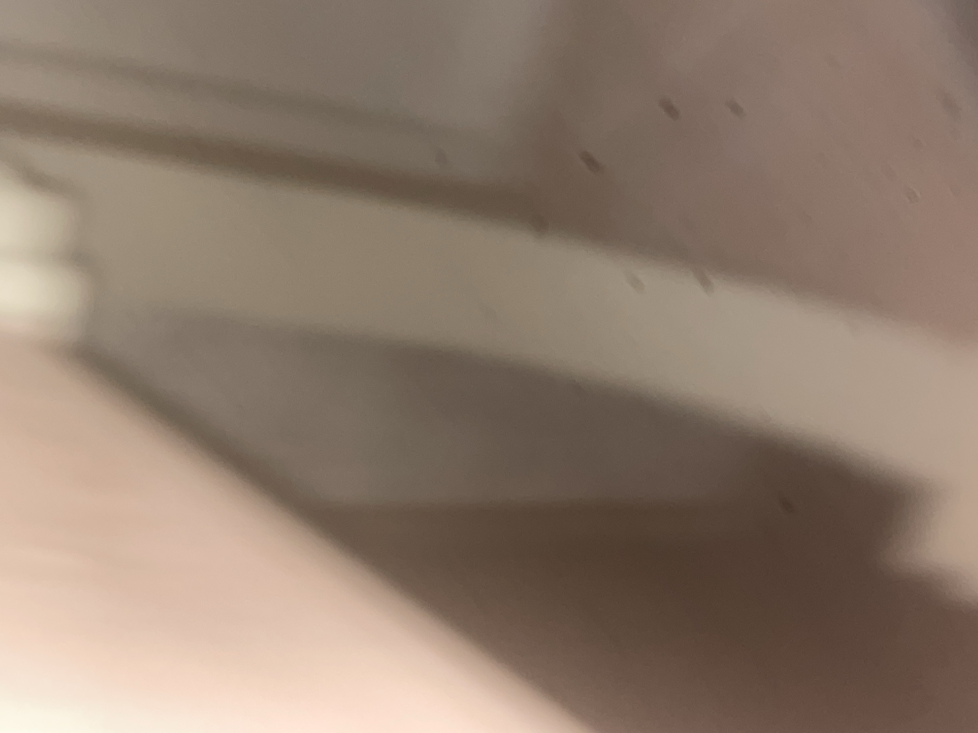 This is a close-up, out-of-focus photograph of a wall and a vertical element, possibly a door frame or a partition, in Howayi, Nigeria. The wall appears to be a light, peachy color, and there are subtle variations in shading that suggest lighting from the upper left. The vertical element is a darker, neutral tone, creating a distinct line of division in the frame. The surface of the wall and the vertical element exhibits numerous small, dark specks, which could be dust, imperfections in the surface, or minor damage. No people or specific objects are clearly discernible due to the lack of focus. The environment appears to be indoors, likely a residential or commercial building, but no specific details about its function are available.