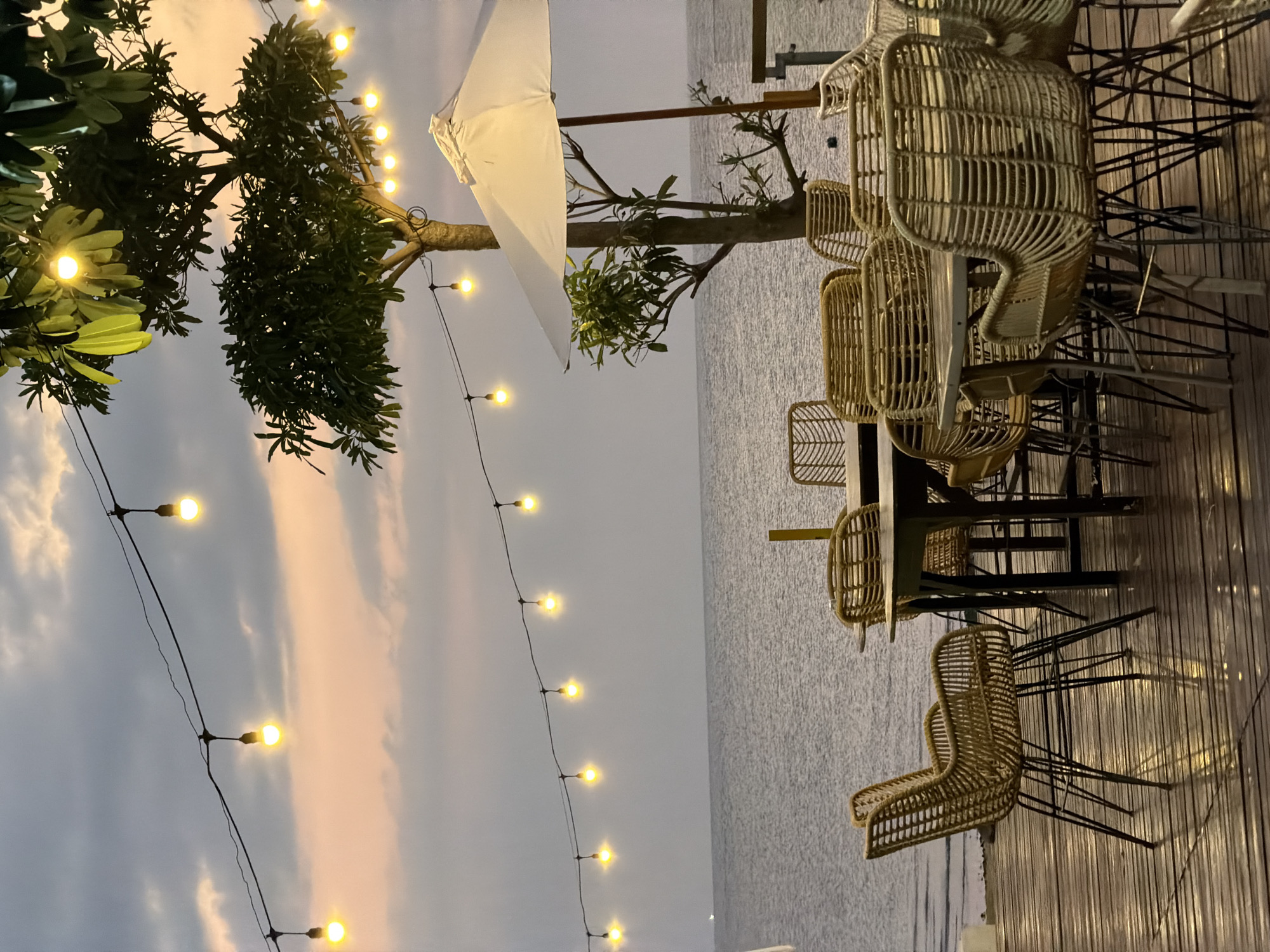 This image captures an outdoor seating area at dusk, likely at a beachfront restaurant or cafe in Pasir Putih, Indonesia. The scene is set on a wooden deck overlooking the ocean, with the water stretching out to the horizon under a cloudy sky that is illuminated with soft hues of pink and orange from the setting sun.
The primary subjects are the rattan chairs and tables, arranged to face the sea. Several chairs are visible, some clustered around tables, while others are positioned individually. The chairs have a distinctive woven design, giving the area a relaxed, tropical ambiance. Overhead, strings of warm-toned Edison bulbs are strung across, casting a gentle glow and enhancing the evening atmosphere. A white umbrella, possibly for shade during the day, is also partially visible.
The time of day is clearly evening, as indicated by the soft, diffused light and the colorful sky. The weather appears to be overcast, but not stormy, with clouds creating a textured backdrop. The presence of the ocean and the tropical foliage suggests a coastal location. The overall mood conveyed is one of tranquility and relaxation, ideal for enjoying a quiet evening by the sea. No people are visible in the image, allowing the focus to remain on the serene setting and the thoughtfully arranged furniture. There is no visible text in the image.