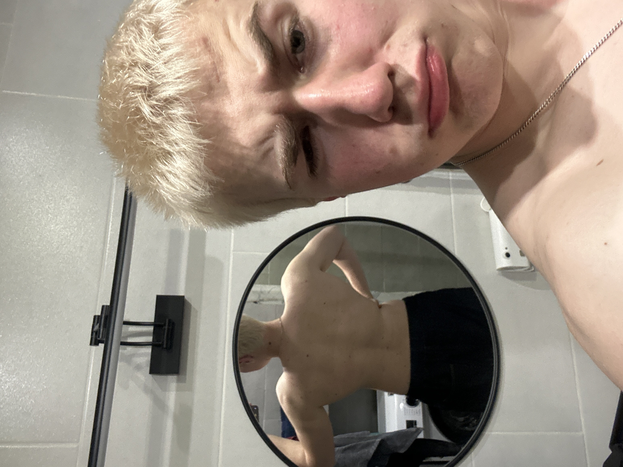 An image captures a close-up, tilted, and high-angle selfie of a light-skinned individual with short, bleached blonde hair. The person's face, featuring a slightly pouty expression and one eye partially closed, dominates the upper part of the frame. They are wearing a thin silver chain necklace around their neck.
Directly below and to the right, a round, black-framed mirror reflects the person's shirtless back, broad shoulders, and arms. The individual in the mirror is seen from behind, angled slightly, showcasing their musculature and wearing dark shorts or underwear.
The background of the main image and the reflection reveals light-colored, possibly tiled, walls, suggesting an indoor setting like a bathroom or a utilitarian space. On the left wall, a black vertical bar with a horizontal support is visible. On the right wall, a white electrical outlet or switch is mounted. Within the mirror's reflection, a dark grey or black towel is draped over what appears to be an appliance, possibly a washing machine, though no clear text is legible on it. The overall lighting is artificial and uniform, offering no clues about the time of day. The person's facial expression conveys a hint of playfulness or a casual pose for the selfie.