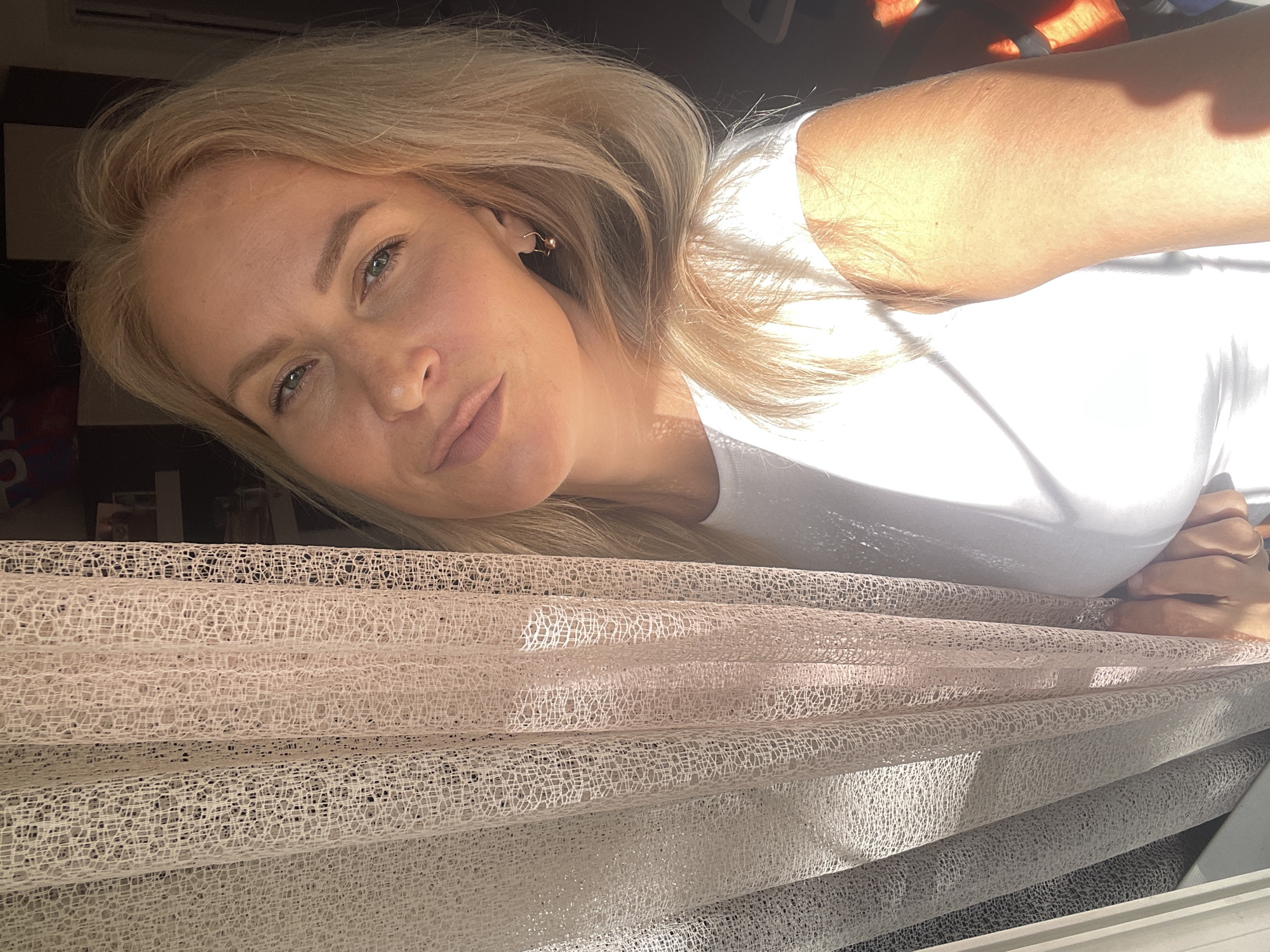A fair-skinned, blonde woman is the primary subject of this image. She has light eyes and wears a white t-shirt. Her face is angled towards the camera with a slight smile. She wears earrings.
In the foreground, there are stacked rolls of fabric, possibly tulle or netting, with a variety of pastel shades.
The lighting is bright and sunny. The location and setting are unknown, and there is nothing in the image to determine that the location is Sochi, Russia.