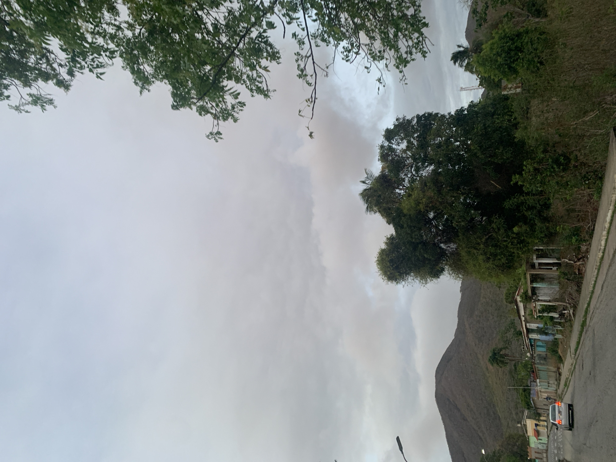 The image depicts a mountainous landscape under an overcast sky, with a road running through a semi-urban area. Green tree branches with leaves form a canopy across the top of the frame, partially obscuring the sky. The sky is entirely covered in dense, gray clouds, suggesting an overcast day with muted light.
A paved road stretches across the lower part of the scene. To the right of the road, a steep hillside rises, covered in a mix of dense green foliage and drier, sparser vegetation. Further in the background, a large, dark mountain dominates the right side of the image, showcasing its rugged terrain.
Along the right side of the road, at the base of the hillside, several small, simple structures are visible. These appear to be houses or shacks, some with brightly painted walls in shades of blue, yellow, and green, and corrugated metal roofs. One structure looks like an abandoned or unfinished building with exposed framing. A white van, with its red taillights illuminated, is visible on the road, heading away from the viewer. Several streetlights are also visible along the road. High on the hillside, amidst the trees, a tall, white pole or antenna structure can be seen. No people are present in the image. The scene conveys a quiet, somewhat somber atmosphere due to the heavy cloud cover.