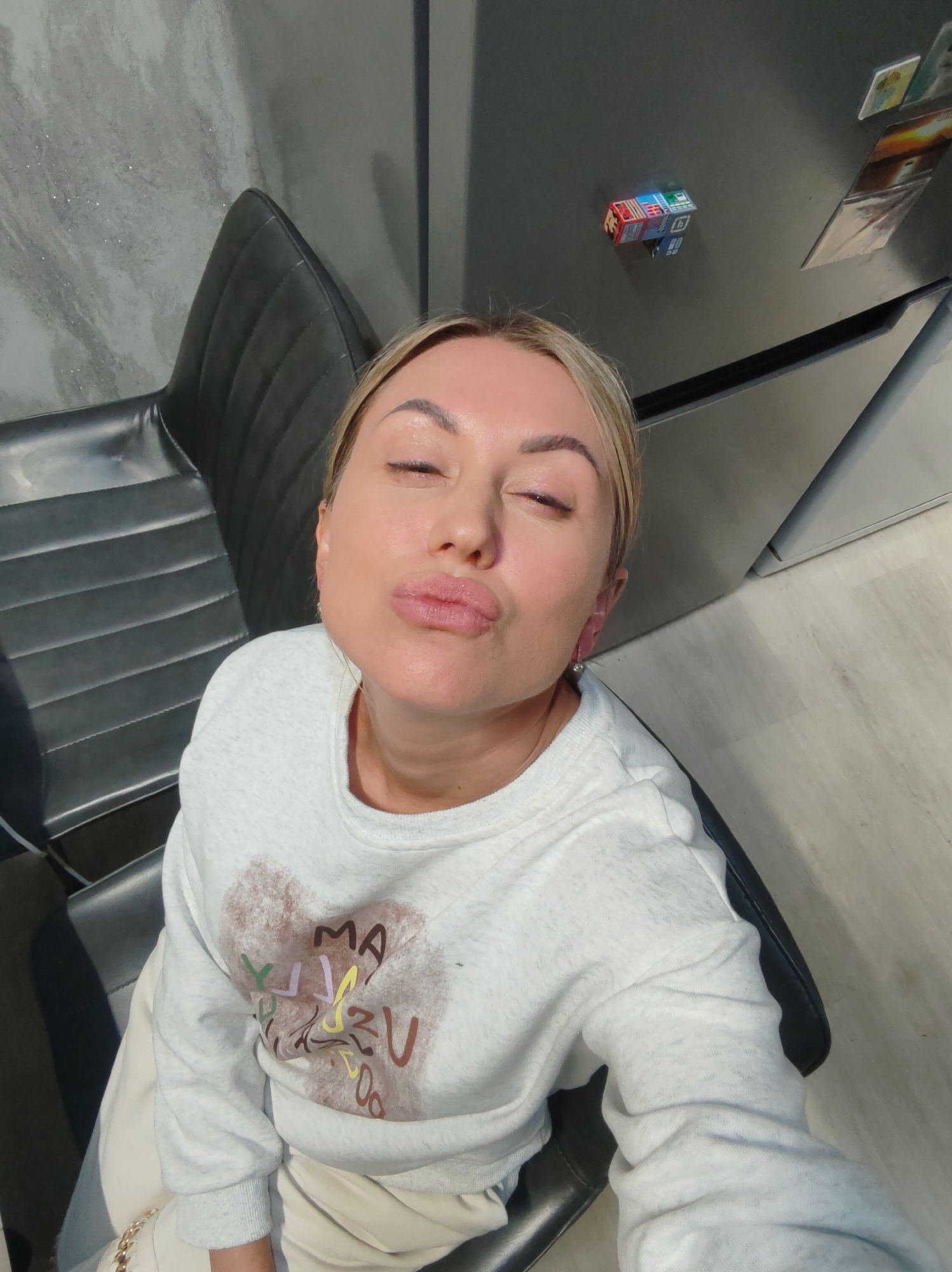 A woman with blonde hair and closed eyes puckers her lips for a selfie in what appears to be an indoor setting. She is wearing a light gray sweatshirt with a graphic design on the front. She is seated in a black chair with a textured seat. In the background, a gray marble-patterned wall is visible, as is a refrigerator with magnets on it. The floor is a light-colored wood or laminate. The overall impression is of a casual, personal moment captured indoors. The location is stated to be Komsomolsk-on-Amur, Russia.