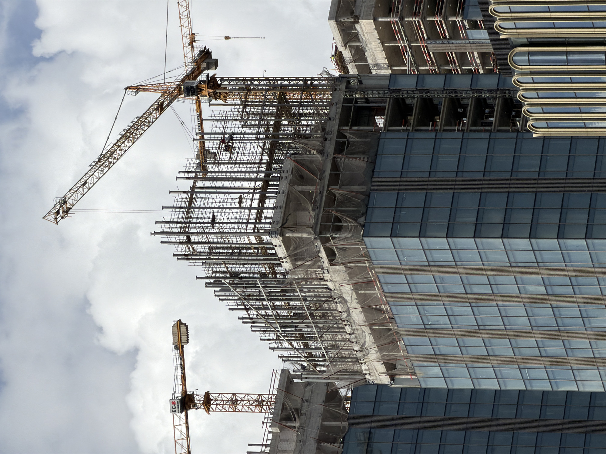 The image shows a high-rise building under construction. Scaffolding surrounds the unfinished sections, and two yellow tower cranes are visible, one extending upwards and the other standing adjacent to the building. The completed section of the building features a glass facade with a grid-like pattern. The sky is filled with clouds, giving a sense of the height of the structure. The building is being constructed in Moscow, Russia.