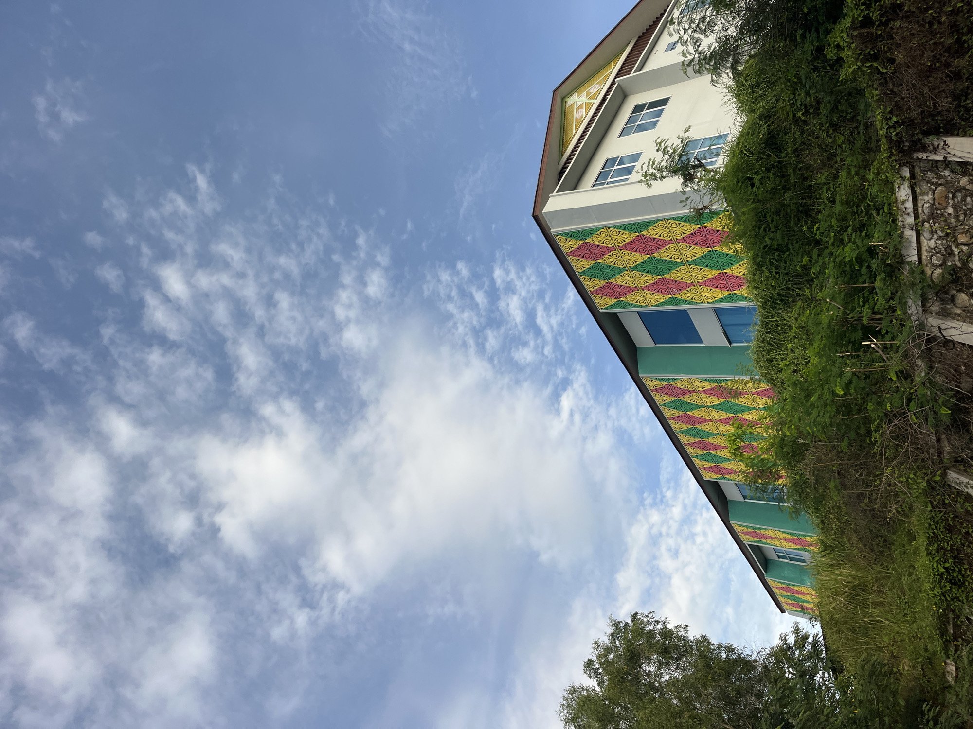 The image depicts a building in Lhokseumawe, Indonesia, viewed from a low angle. The building has a white facade with sections adorned with a yellow, red, and green diamond pattern. Windows are visible along the building's facade. Part of the building is covered with dense green foliage. Above the building is a bright blue sky with scattered white clouds. Trees are visible at the bottom of the frame.