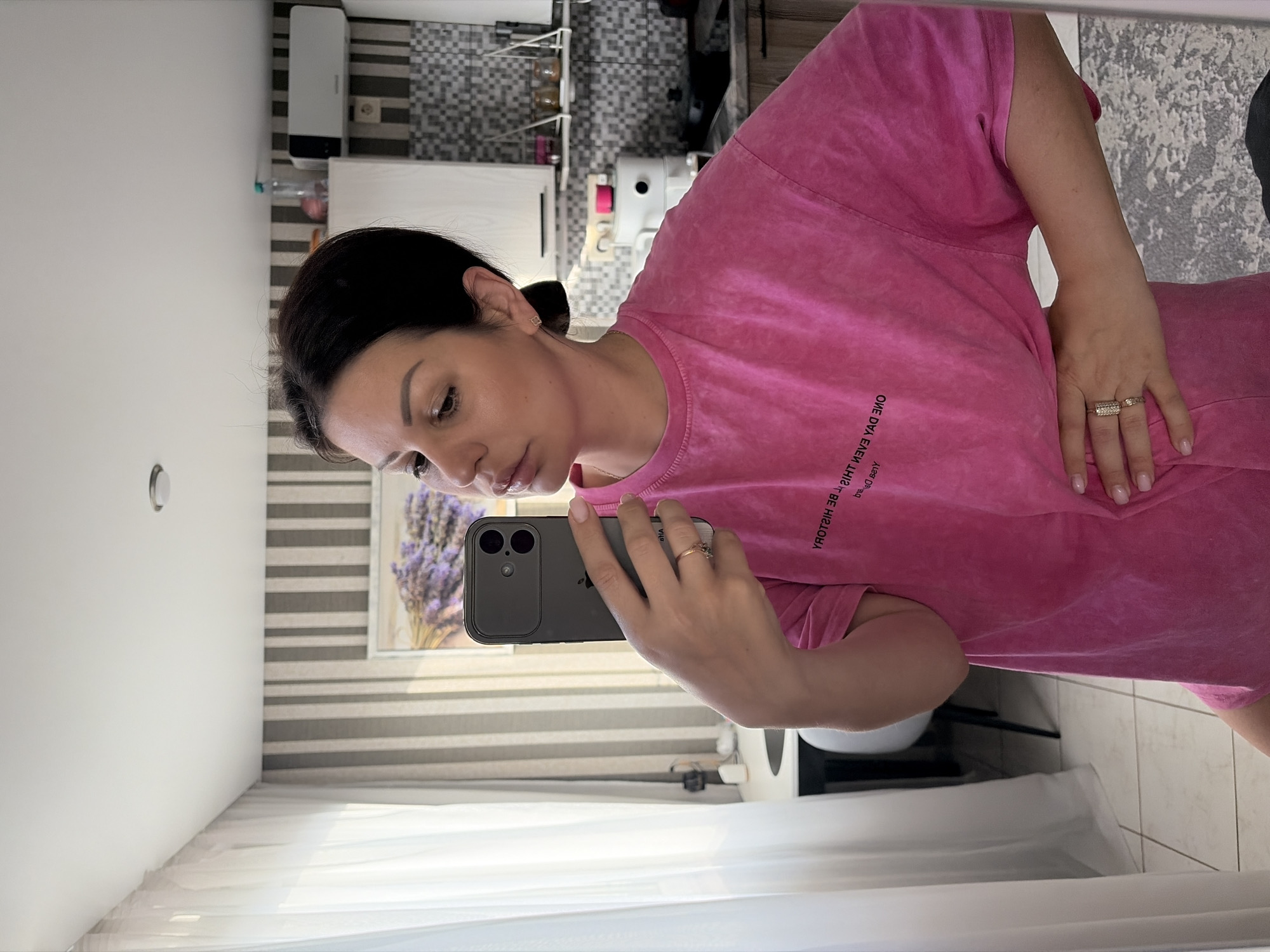 A woman with dark hair pulled back in a bun is taking a mirror selfie in what appears to be a kitchen area. She is wearing a bright pink t-shirt with text printed on it in black. Her left hand is holding a smartphone up to take the picture, and her right hand is resting on her hip. The background shows a tiled floor, vertical striped wallpaper, a window with white curtains, kitchen cabinets, a shelf with jars, and a white air conditioning unit. A framed picture of lavender is visible on the wall. The overall scene suggests an indoor residential setting. The specific location within Sochi, Russia, cannot be determined from the visual information provided.