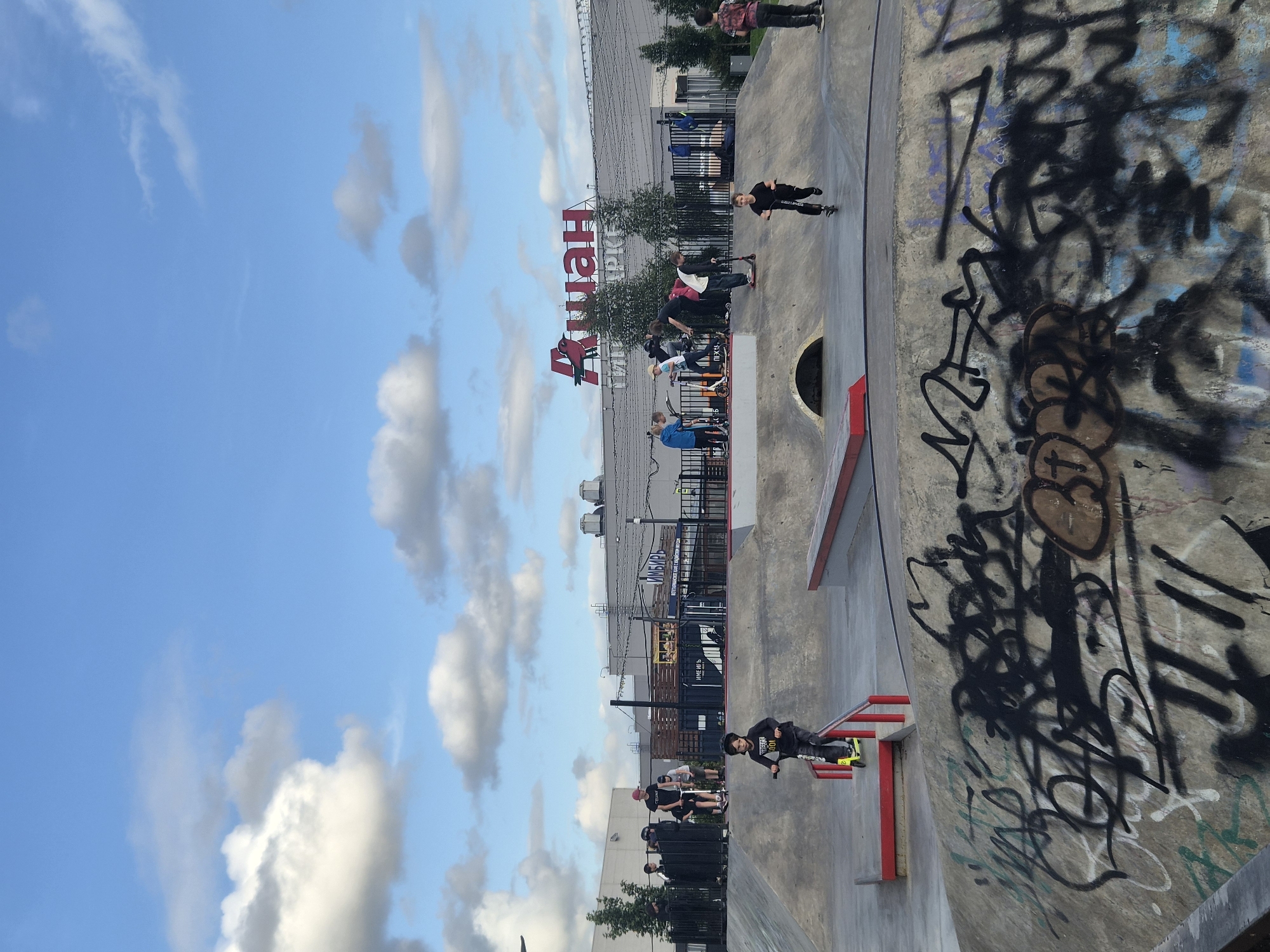 This is a vertical, eye-level, medium-wide shot of a skatepark in Kudrovo, Russia, under a bright blue sky with scattered clouds. In the foreground, a young boy on a scooter rides past a metal rail. The concrete skatepark is covered in graffiti. In the middle ground, several other children are on scooters in various parts of the skatepark. Behind the skatepark, a black metal fence encloses the area. Beyond the fence, a large building with a red logo that reads "АШАН" (Auchan) is visible, along with trees and smaller structures. The overall scene depicts recreational activity on a pleasant day.