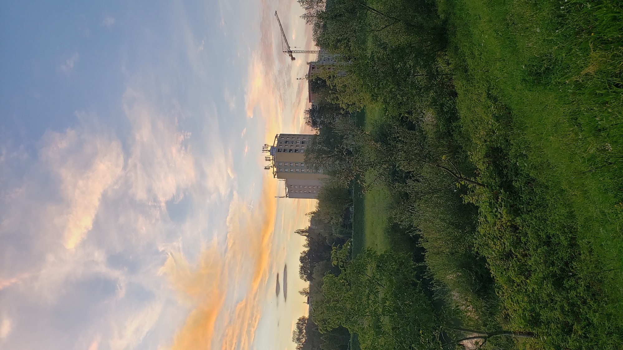 The image depicts a view of the city of Prešov, Slovakia, likely from a green space or park overlooking the area. In the background, a tall, light-colored apartment building with multiple floors is visible, topped with several antennas or communication dishes. To the right of this building, a construction crane is present, suggesting ongoing development or renovation.
The sky above is a gradient of blue, transitioning into hues of soft orange and pink from the setting or rising sun, with scattered clouds adding texture. The middle ground features a band of trees and vegetation, separating the sky from the foreground.
The foreground is dominated by lush green grass and various types of shrubbery and trees, suggesting a natural or semi-natural environment. The overall impression is one of urban scenery blended with natural elements, captured during twilight. The specific location within Prešov cannot be determined solely from the visual information provided.